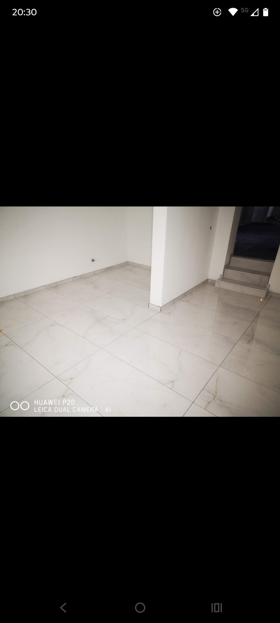 Jasne, marmurowe płytki podłogowe w pustym pomieszczeniu z białymi ścianami i schodami w tle. Widoczny fragment instalacji na ścianie.