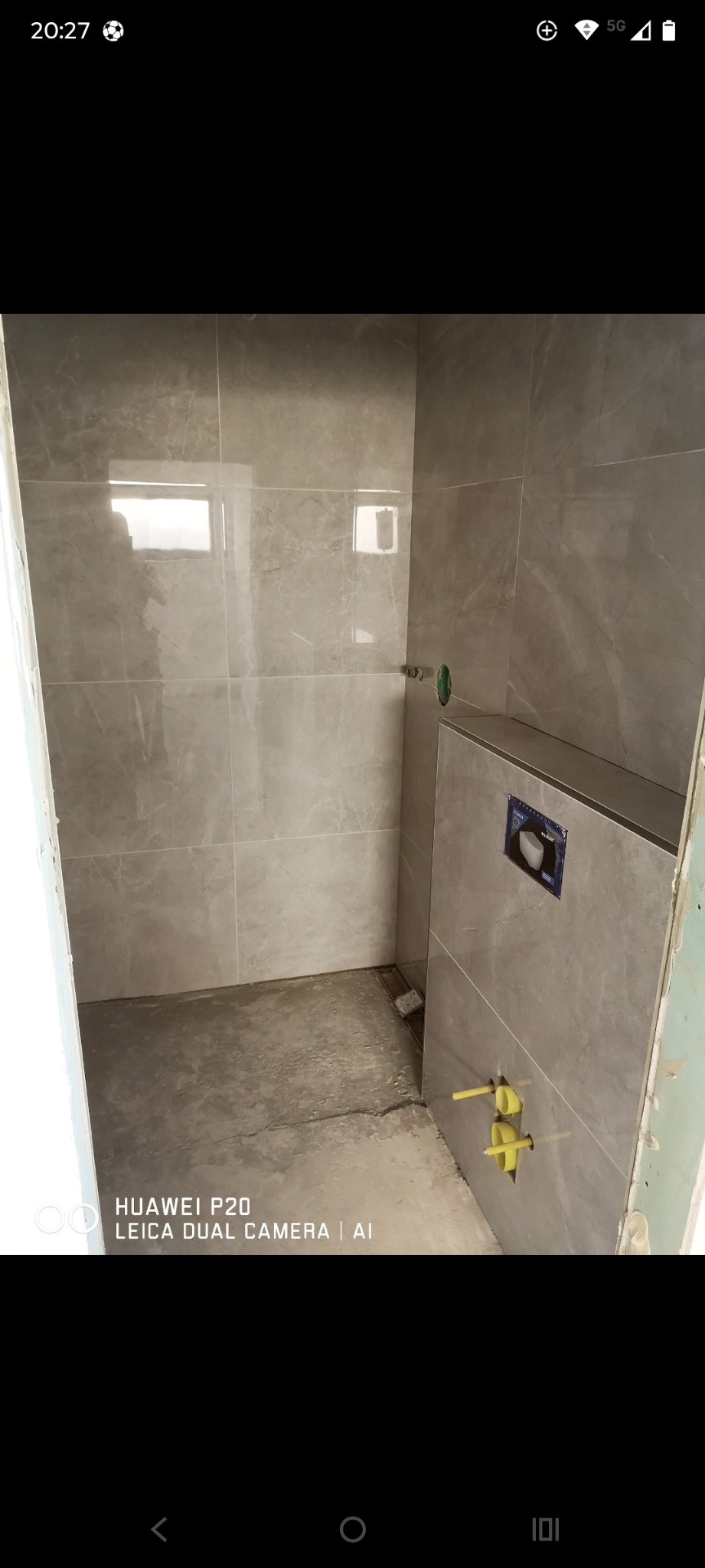 Wykończona łazienka z szarymi płytkami imitującymi marmur, widoczne elementy instalacji wodnej i odpływ liniowy. Płytki ułożone do połowy wysokości ściany.