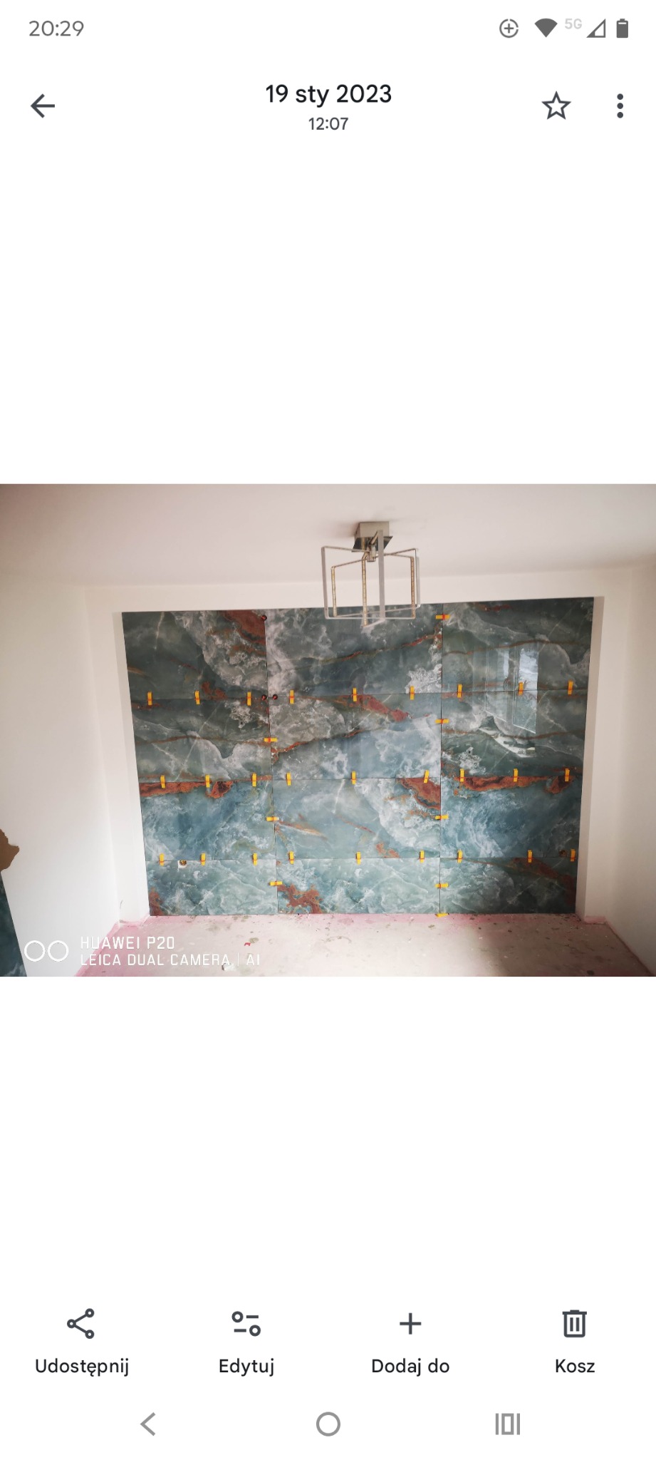 Ściana w trakcie układania płytek imitujących marmur, z użyciem żółtych krzyżyków dystansowych. Widoczna lampa sufitowa i fragment podłogi.