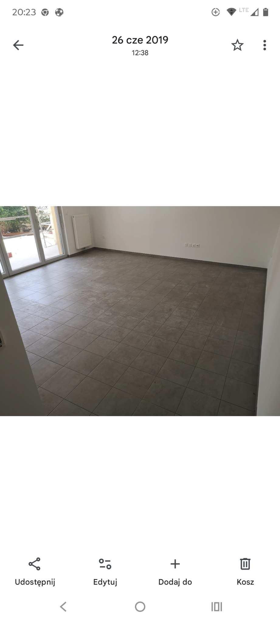 Wykończona podłoga z szarych, kwadratowych płytek w jasnym pokoju z białymi ścianami i dużym oknem balkonowym. Widoczne gniazdka elektryczne przy ścianie.