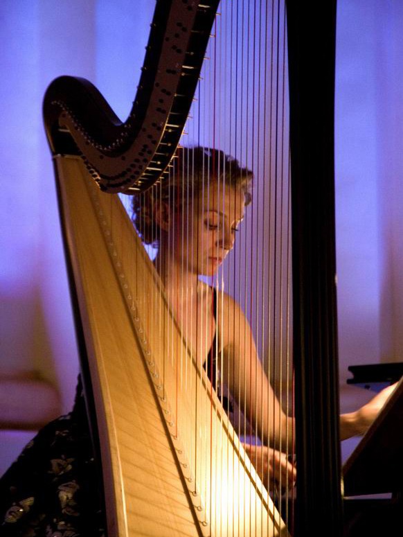 Elegancka harfistka w skupieniu gra na harfie koncertowej, oświetlona subtelnym, fioletowym światłem scenicznym. Widoczne detale instrumentu i strój artystki.