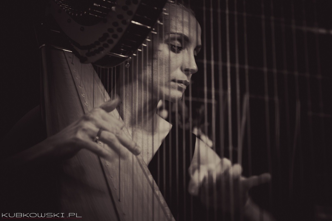 Artystyczne, monochromatyczne ujęcie harfistki w trakcie gry. Widoczne dłonie na strunach, fragment harfy i profil kobiety. Delikatne światło i głębia ostrości.