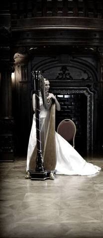 Elegancka harfistka w białej sukni, prezentująca instrument na tle ciemnego, zdobionego wnętrza. Ujęcie podkreśla artystyczny charakter występu.