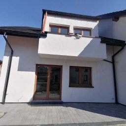 Jozef Zablotny - Nowoczesny dom z białym tynkiem, ciemną stolarką okienną i dachem. Widoczny fragment tarasu z szarej kostki brukowej. W tle budynek w trakcie budowy.
