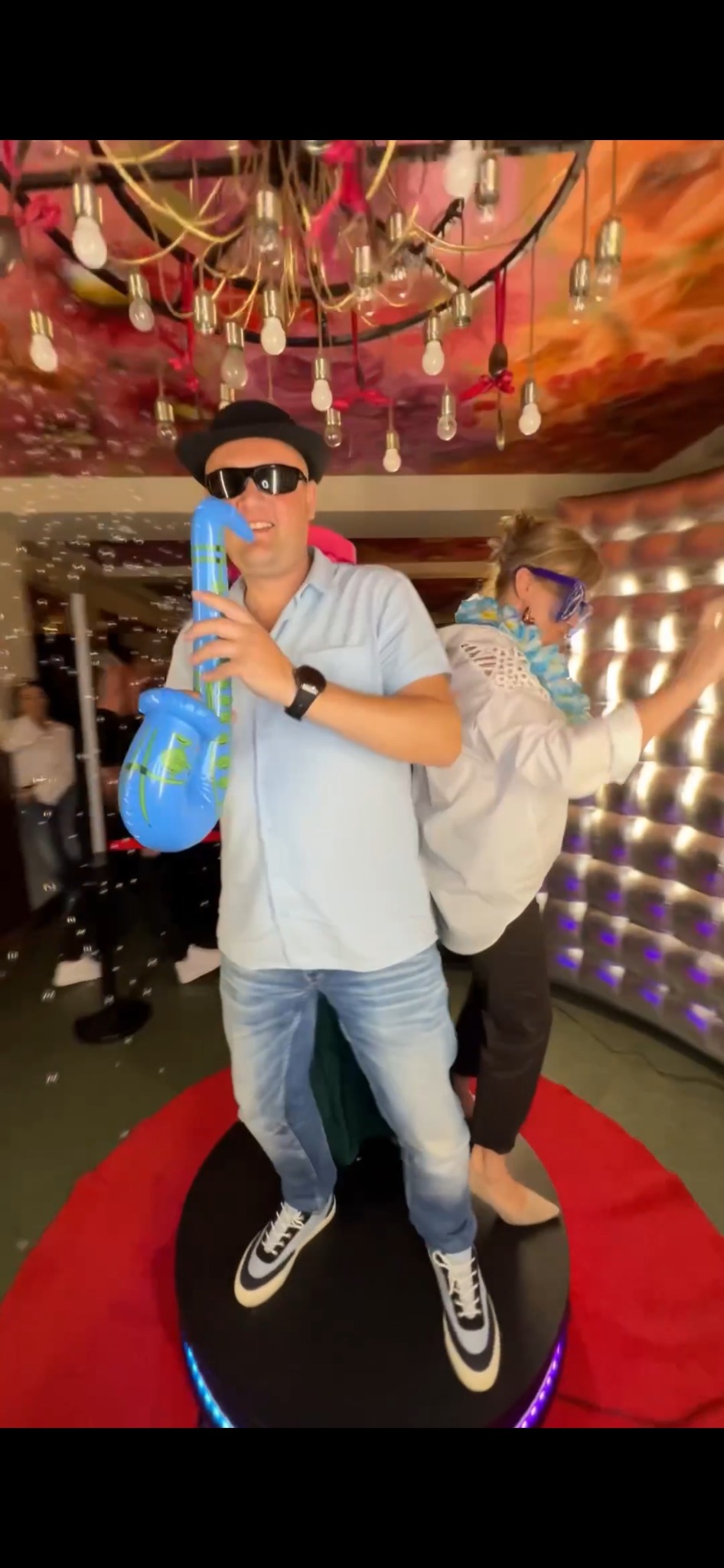 Radosne ujęcie z imprezy: mężczyzna w kapeluszu z dmuchanym saksofonem i kobieta w okularach tańczą na platformie z podświetleniem LED, w tle dekoracyjne oświetlenie.