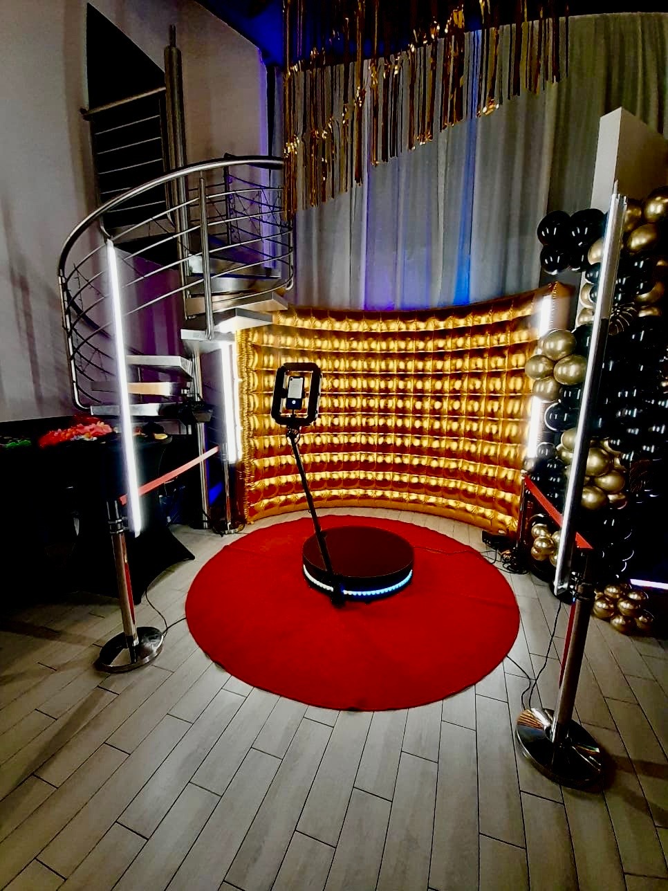 Okrągła platforma z telefonem na statywie, czerwony dywan, złota ścianka i balony w kolorach złota i czerni tworzą elegancką przestrzeń do nagrywania filmów.