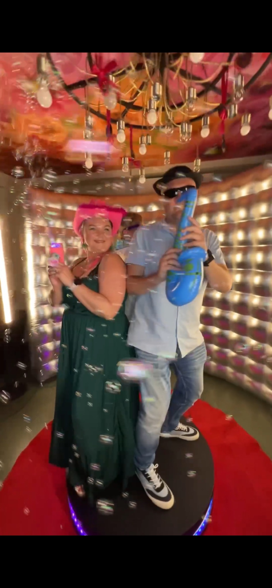 Uśmiechnięta kobieta w różowym kapeluszu i mężczyzna z dmuchaną gitarą pozują wewnątrz błyszczącej, oświetlonej konstrukcji z czerwonym dywanem i bańkami mydlanymi.