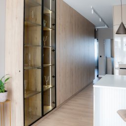 P2Studio Mateusz Grzesik - Nowoczesna zabudowa meblowa z podświetlaną witryną na szkło, drewniane fronty, jasna podłoga i minimalistyczne oświetlenie sufitowe w eleganckim wnętrzu.