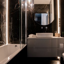 P2Studio Mateusz Grzesik - Nowoczesna łazienka z czarnymi płytkami imitującymi marmur, podświetleniem LED, owalnym lustrem i szarą szafką pod umywalką. Widoczna wanna z parawanem.
