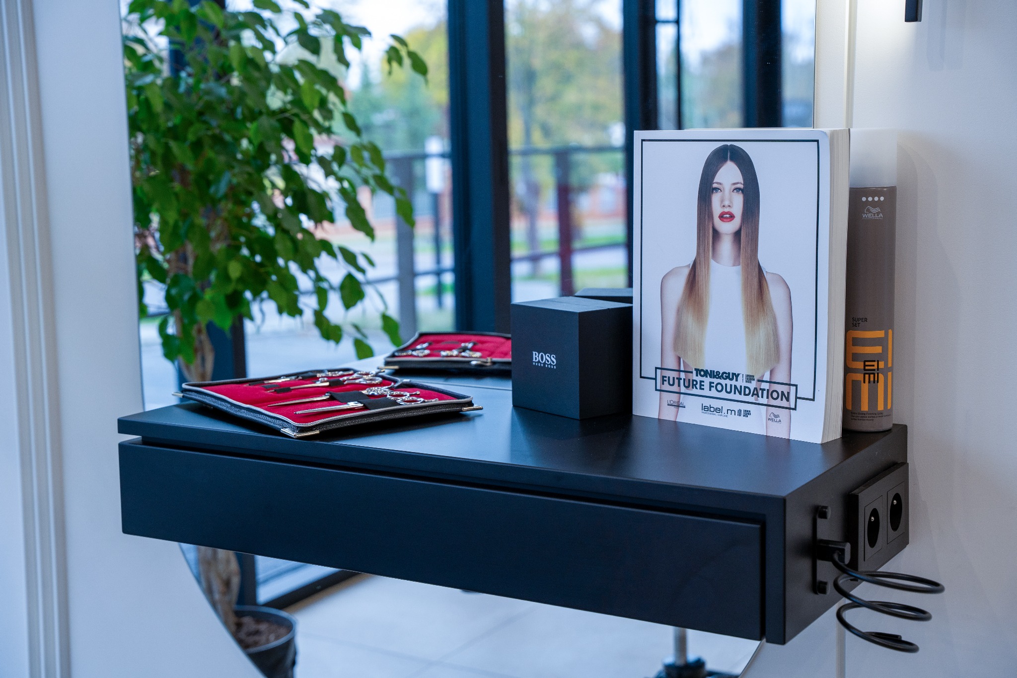 Czarne biurko z akcesoriami: szkatułka z biżuterią, pudełko Hugo Boss, reklama Toni&Guy i kosmetyki Wella. W tle okno i roślina doniczkowa.