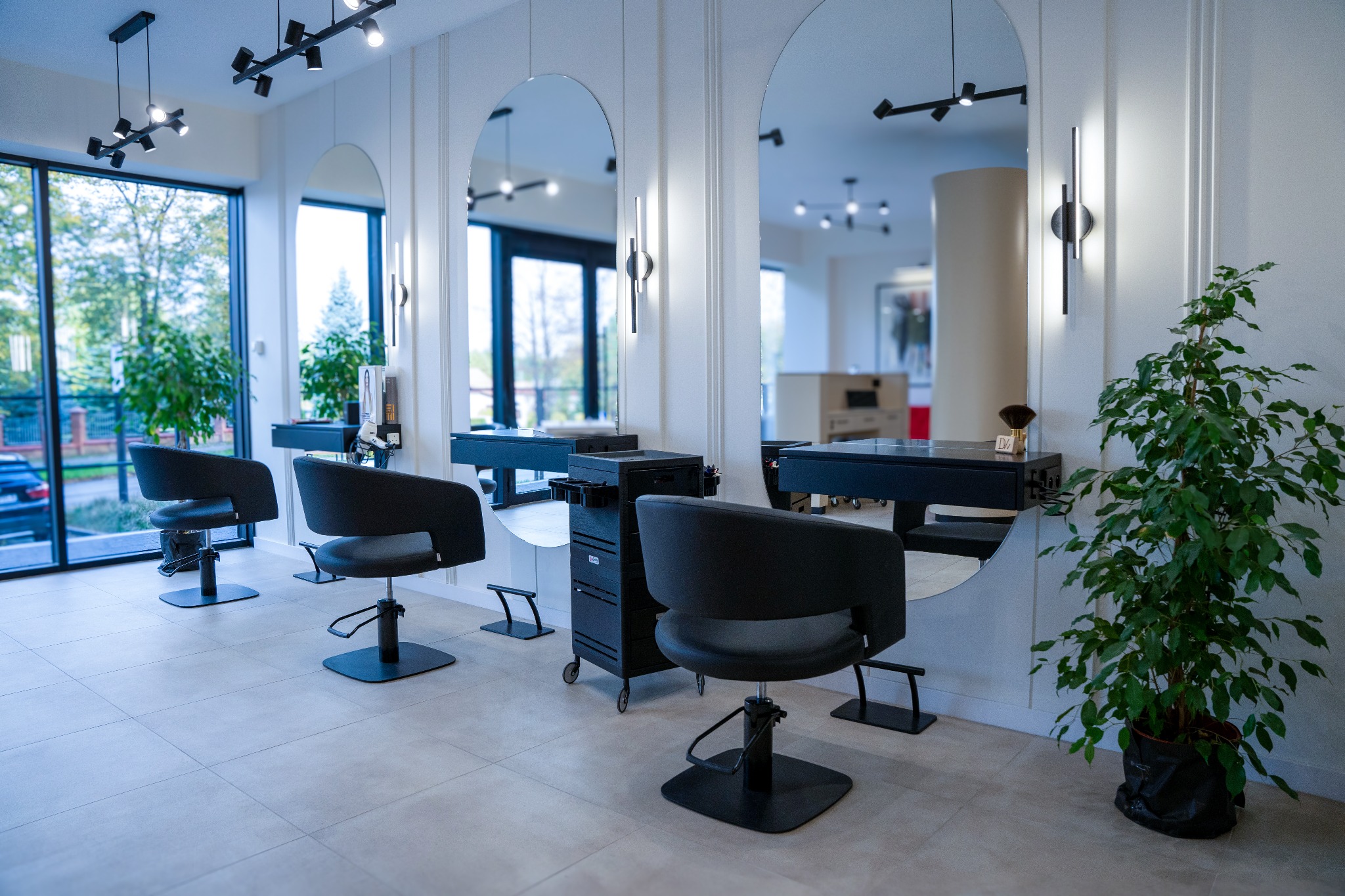 Nowoczesne wnętrze salonu fryzjerskiego z dużymi lustrami, czarnymi fotelami i roślinami doniczkowymi. Jasne płytki podłogowe i oświetlenie punktowe.