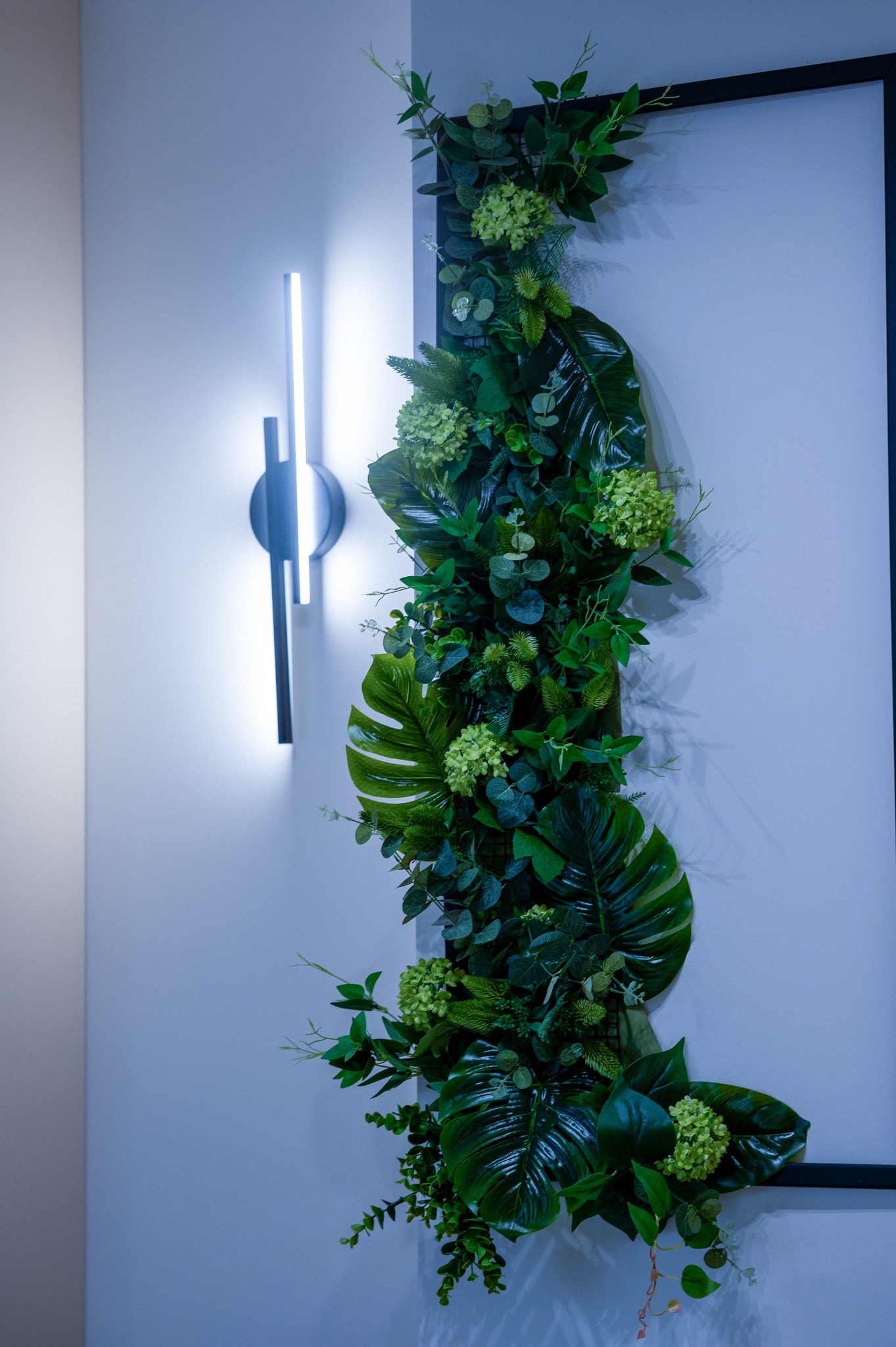 Dekoracja ścienna z zielonych roślin w czarnej ramie, oświetlona nowoczesną lampą LED. Minimalistyczny design, chłodne oświetlenie, gładka ściana w tle.