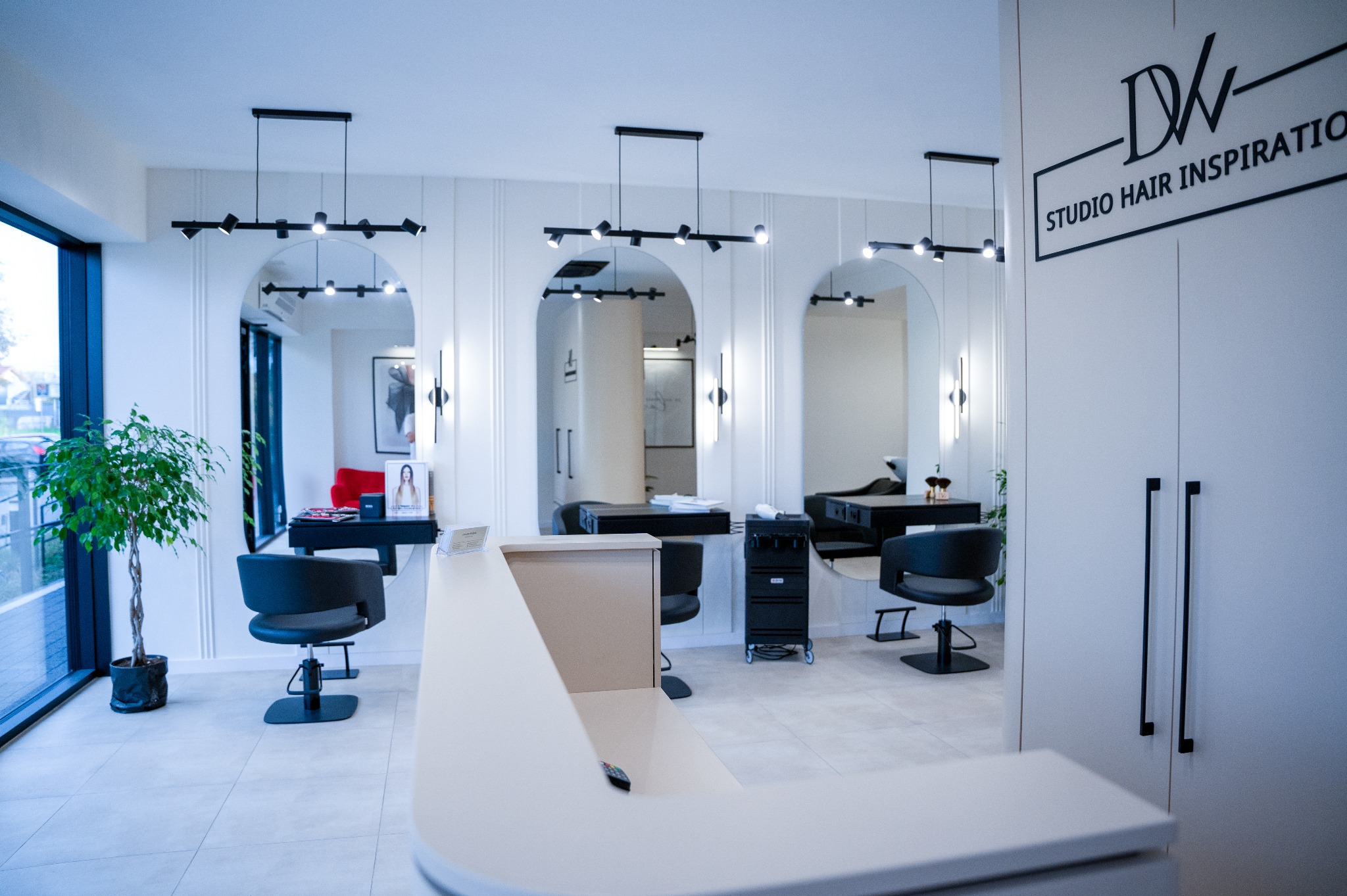 Nowoczesne wnętrze salonu fryzjerskiego z recepcją, stanowiskami pracy z lustrami w łukowych wnękach, oświetleniem punktowym i rośliną doniczkową przy oknie.