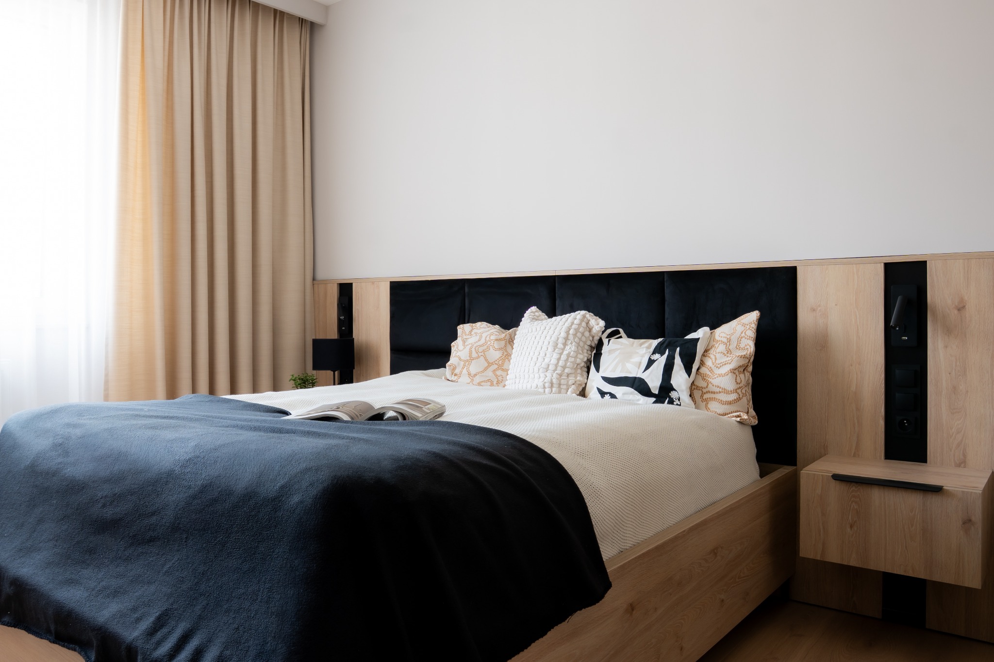 Nowoczesna sypialnia z drewnianym łóżkiem, czarnym zagłówkiem i dekoracyjnymi poduszkami. Na łóżku leży otwarta książka. Stonowana kolorystyka, beżowe zasłony w tle.