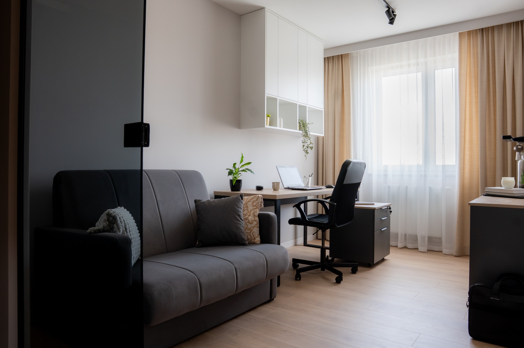 Nowoczesny pokój biurowy z szarą sofą, biurkiem z laptopem i krzesłem biurowym na tle okna z beżowymi zasłonami. Minimalistyczne białe szafki wiszące.