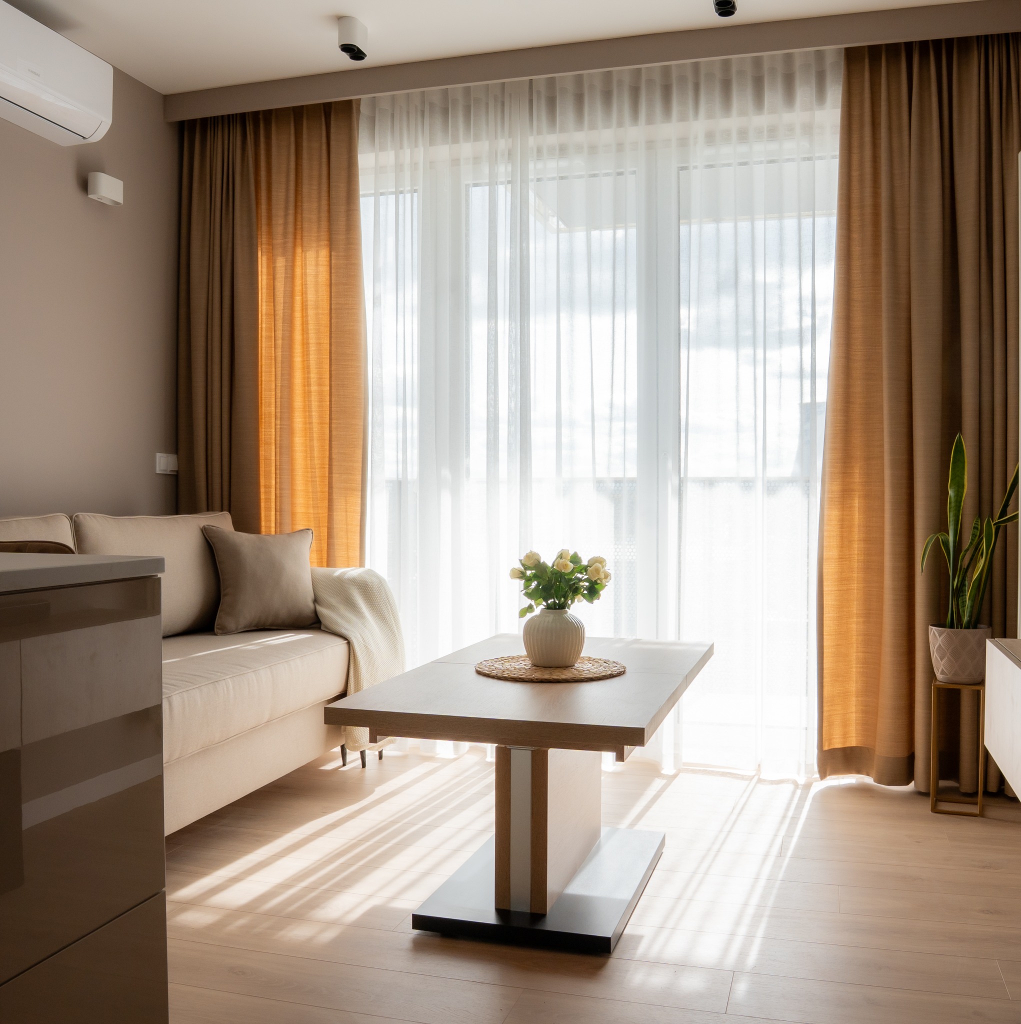 Jasny salon z beżową sofą, drewnianym stołem z kwiatami i promieniami słońca wpadającymi przez okno z brązowymi zasłonami. Minimalistyczny design wnętrza.