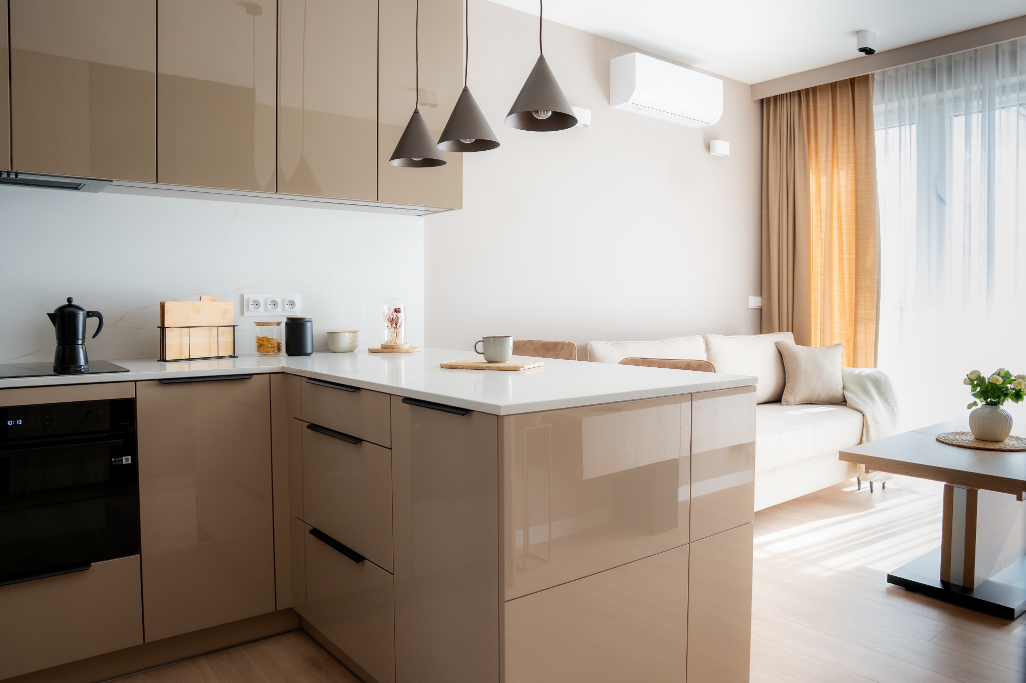 Beżowa, lakierowana kuchnia z wyspą i białym blatem, połączona z salonem. Nad wyspą trzy stożkowe lampy. W tle sofa i okno z jasnymi zasłonami.