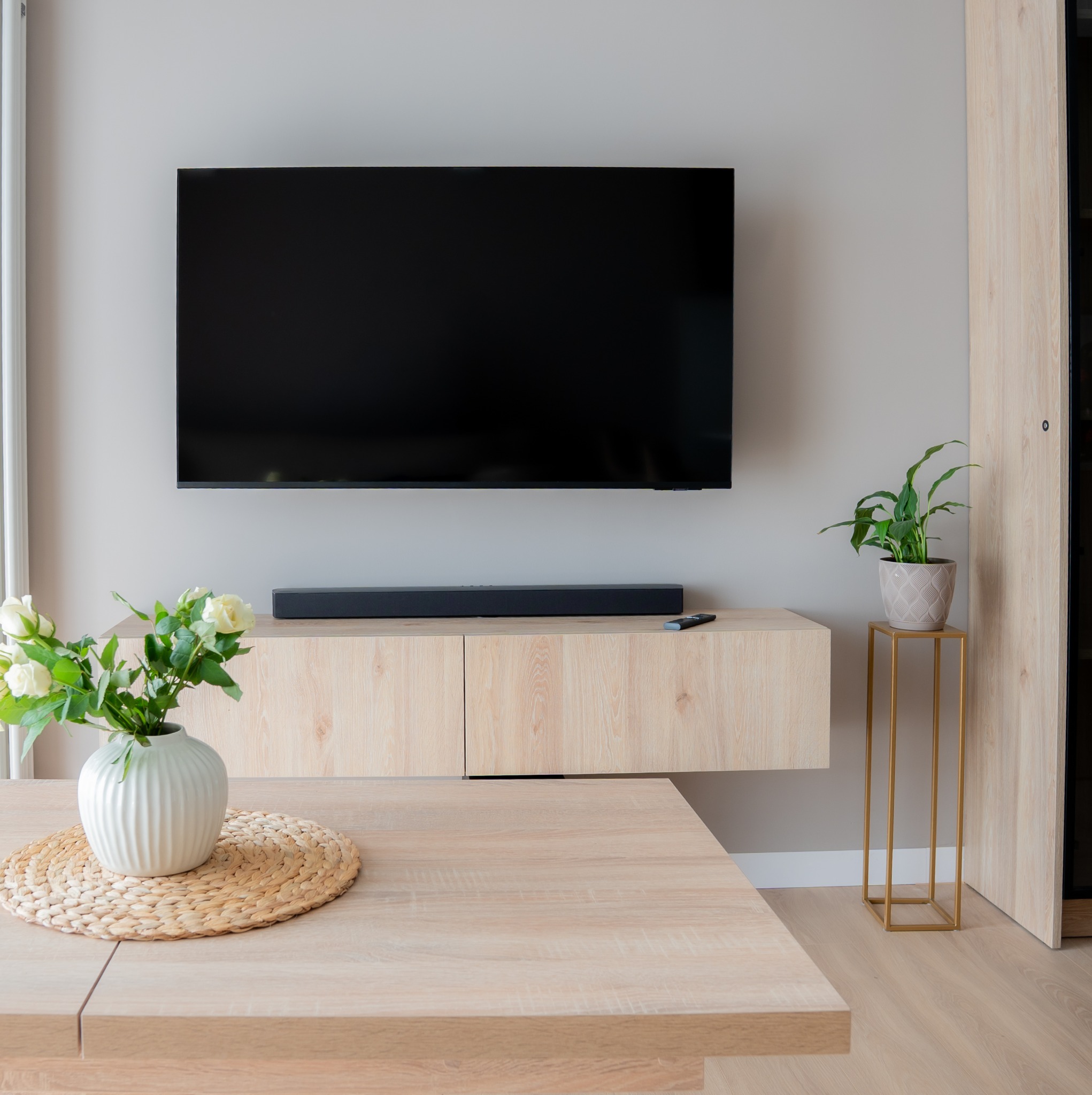 Jasne drewno, minimalistyczny salon: TV na ścianie, soundbar, kwiaty w wazonie na stole, roślina na złotym stojaku. Subtelne, nowoczesne wnętrze.