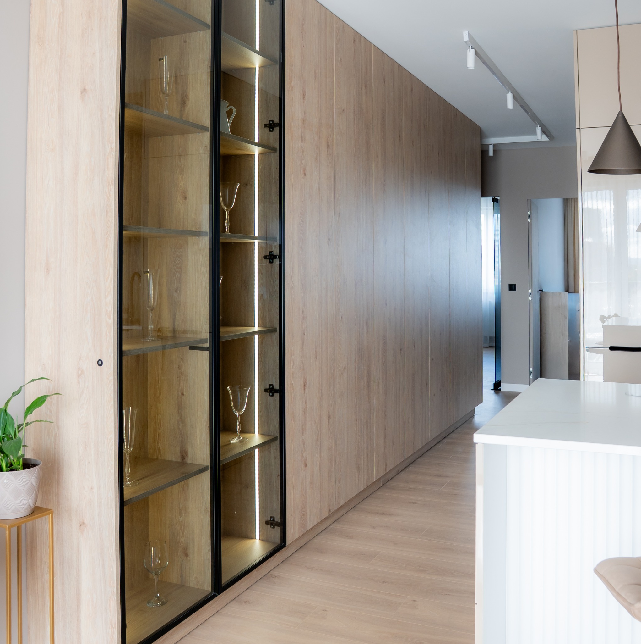 Nowoczesna zabudowa meblowa z podświetlaną witryną na szkło, drewniane fronty, jasna podłoga i minimalistyczne oświetlenie sufitowe w eleganckim wnętrzu.