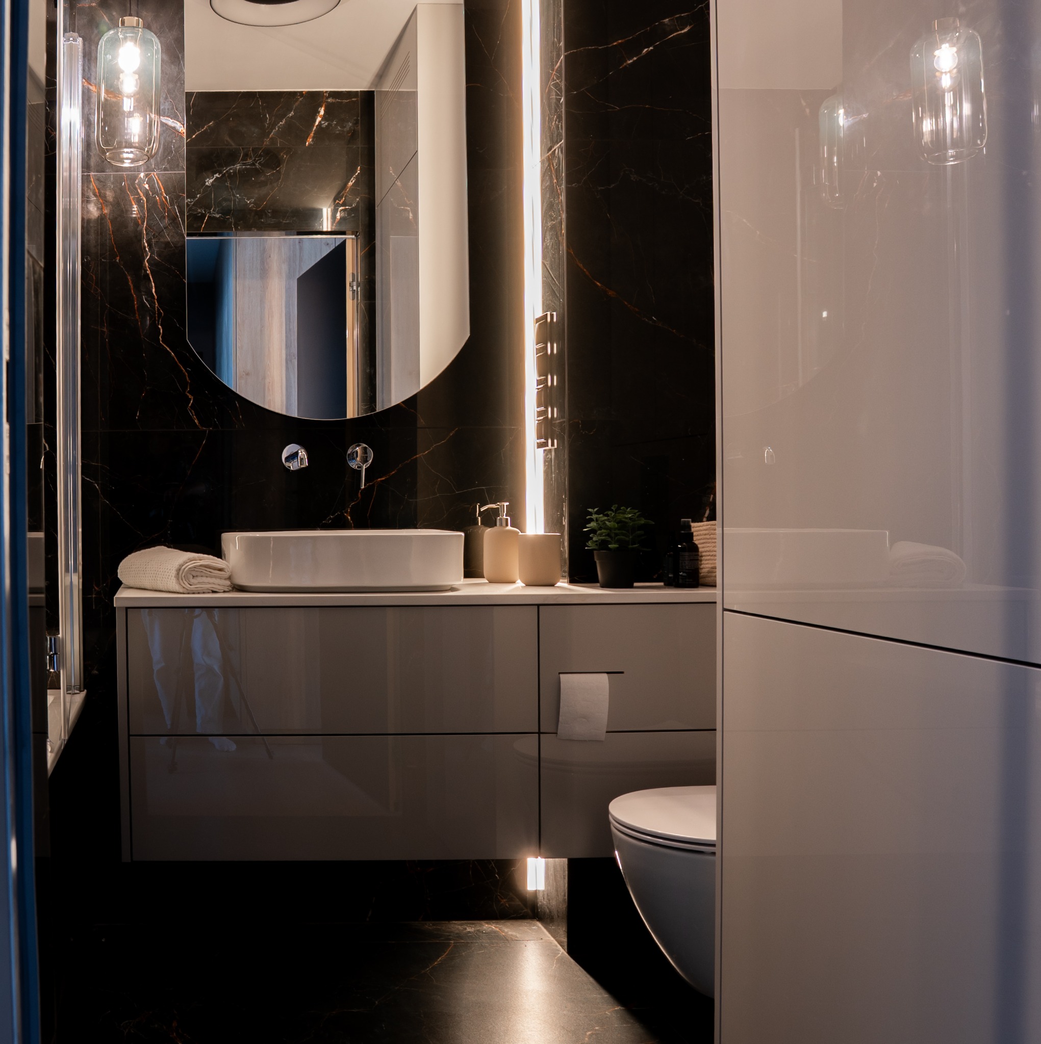 Nowoczesna łazienka z czarnymi marmurowymi płytkami, owalnym lustrem z podświetleniem LED i białą umywalką nablatową. Minimalistyczny design, eleganckie wykończenie.