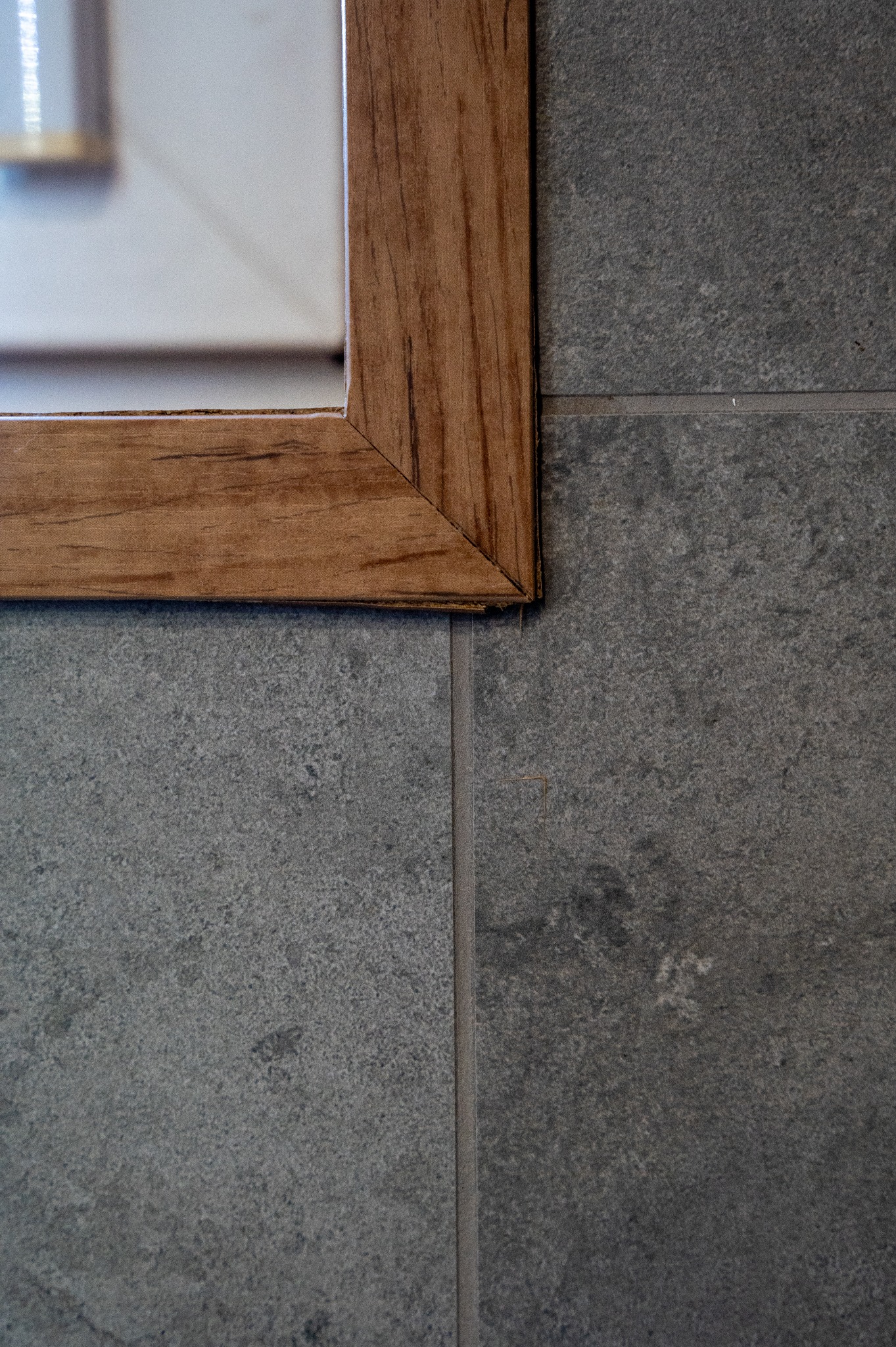 Fragment łazienki: szare płytki gresowe na ścianie, drewniana rama lustra w rogu kadru. Minimalistyczny detal wykończenia wnętrza.