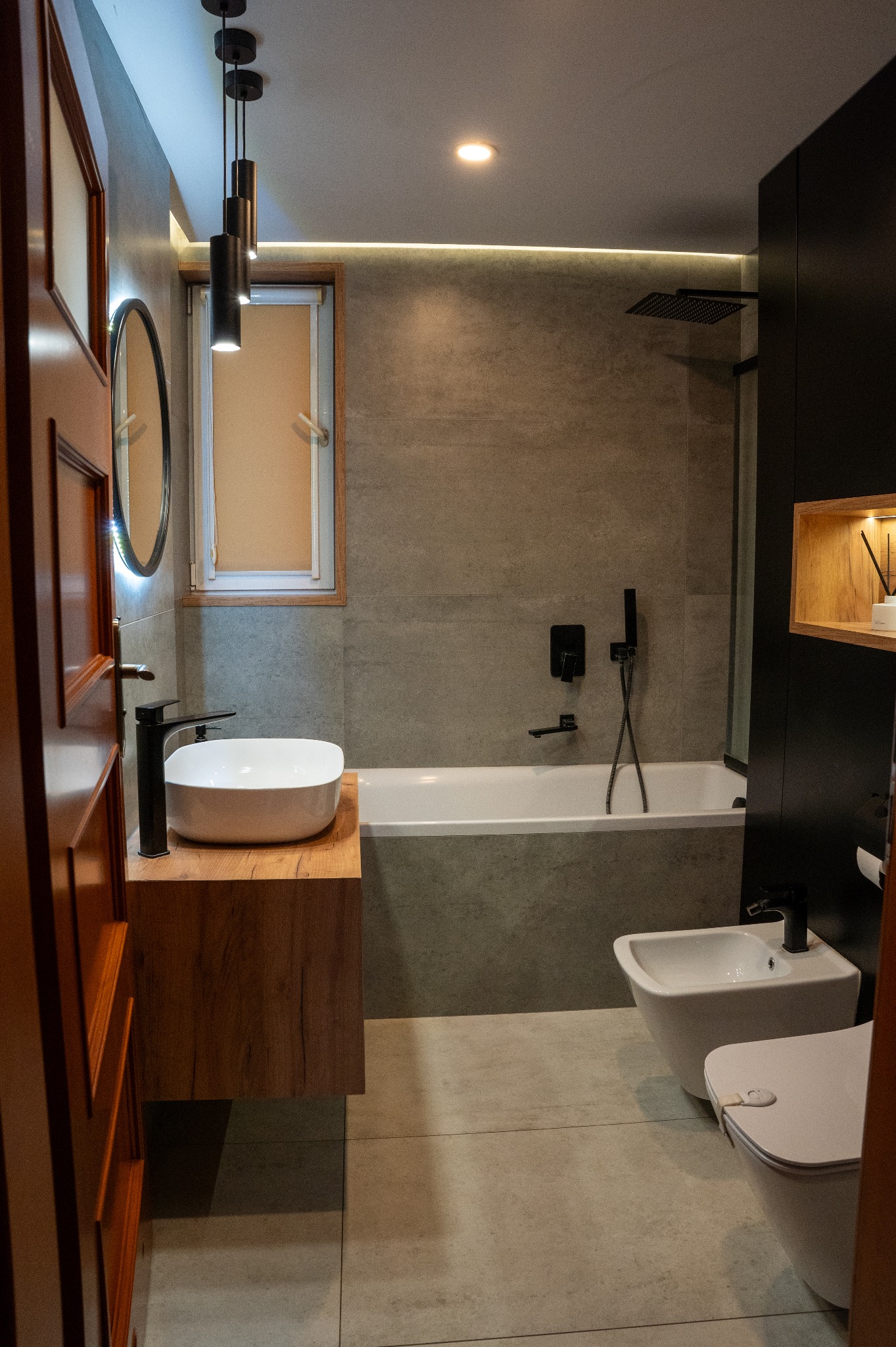 Nowoczesna łazienka z wanną, umywalką nablatową na drewnianej szafce i czarną armaturą. Szare płytki, oświetlenie LED i wiszące lampy dodają elegancji.
