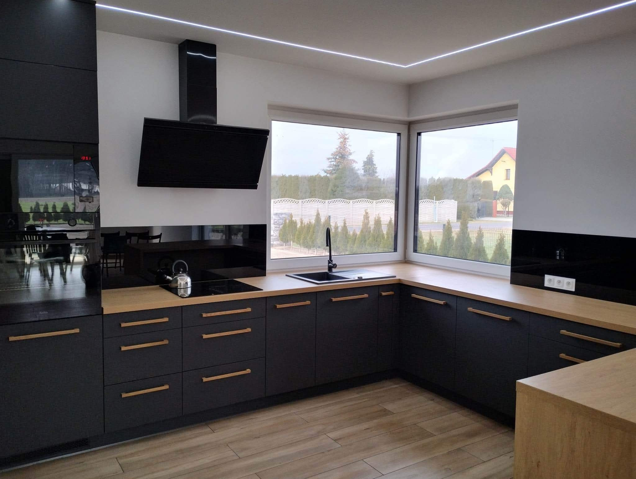 Nowoczesna kuchnia z czarnymi frontami i drewnianymi blatami, oświetleniem LED podszafkowym i dużym oknem z widokiem na ogród. Minimalistyczny design.