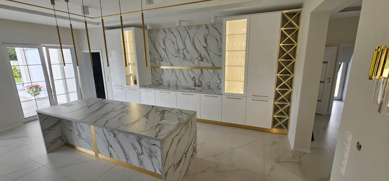 Biała kuchnia z marmurowym blatem i złotymi akcentami. Wyspa kuchenna z marmurowym wzorem. Nowoczesne oświetlenie i eleganckie wykończenie.