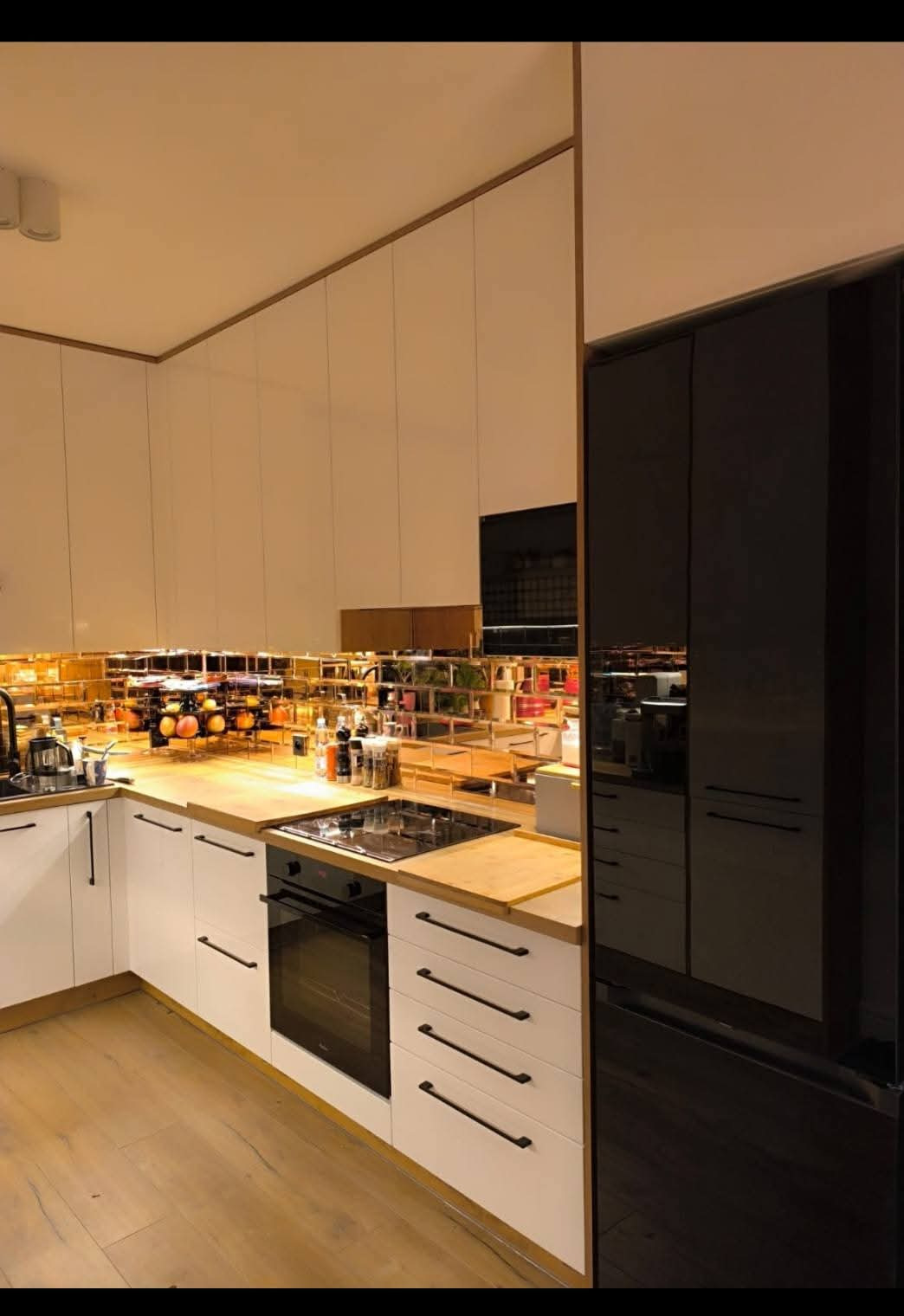 Nowoczesna kuchnia z białymi szafkami, drewnianym blatem i lustrzanym panelem nad blatem, odbijającym akcesoria kuchenne i owoce. Czarna lodówka z połyskiem.