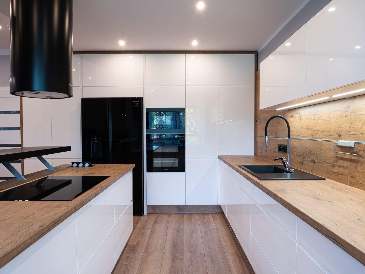 Nowoczesna kuchnia z białymi, lakierowanymi frontami, drewnianym blatem i czarnymi akcentami: okap, zlew i sprzęt AGD. Oświetlenie LED podkreśla ciepło drewna.