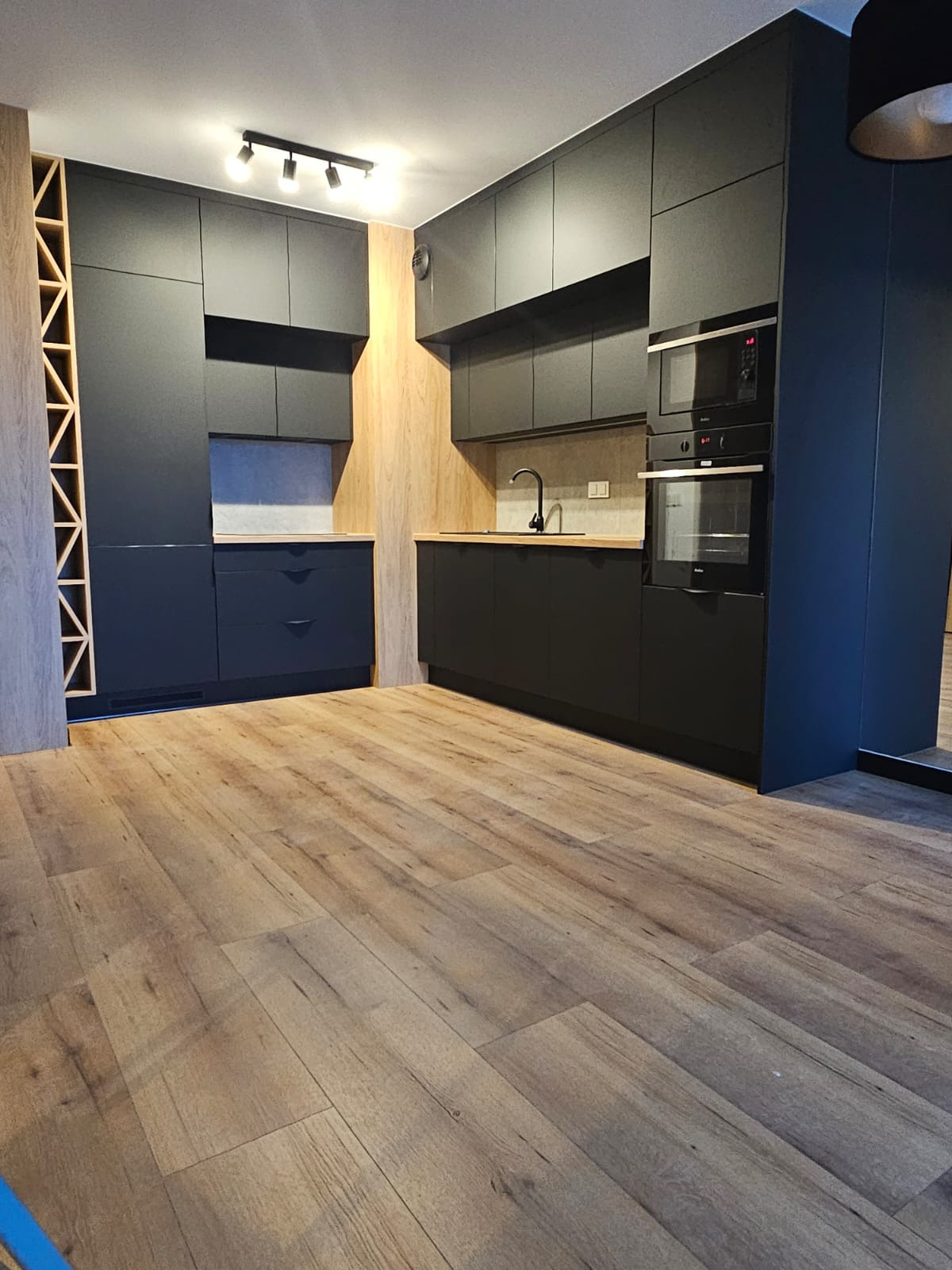 Nowoczesna, czarna zabudowa kuchenna z drewnianymi akcentami i oświetleniem punktowym. Widoczny fragment podłogi z paneli imitujących drewno.