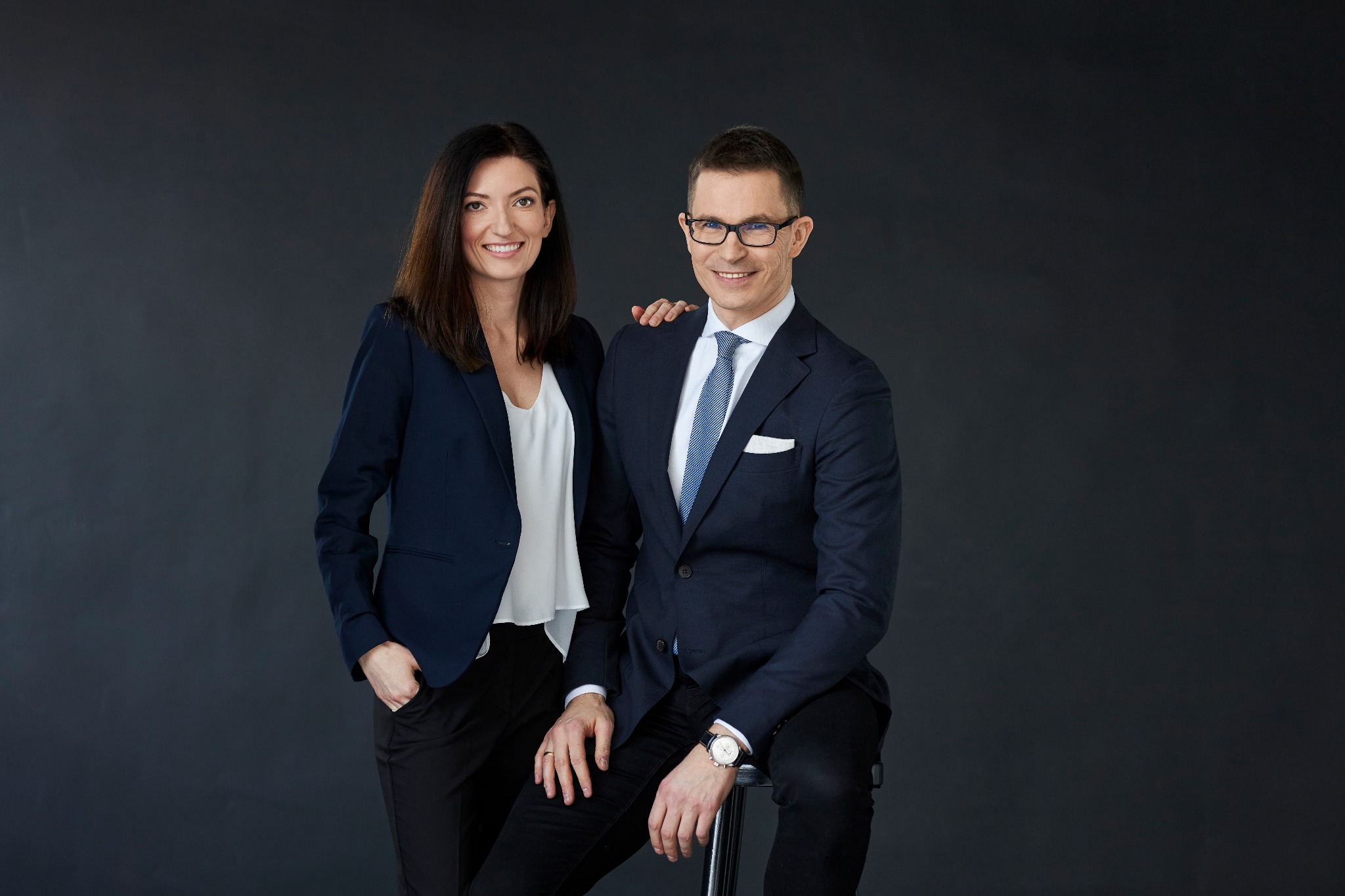 Profesjonalny portret biznesowy: kobieta w granatowej marynarce i mężczyzna w garniturze, oboje uśmiechnięci na ciemnoszarym tle. Mężczyzna siedzi na stołku.