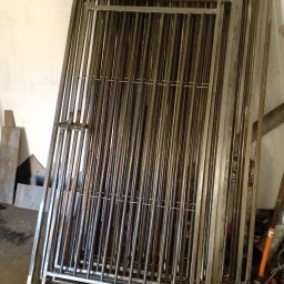 GABJUS Gabriel Feliszczak - Stos metalowych furtek z zamkiem, wykonanych z rurek i profili, opartych o ścianę w warsztacie. Widoczne ślady obróbki i narzędzia.