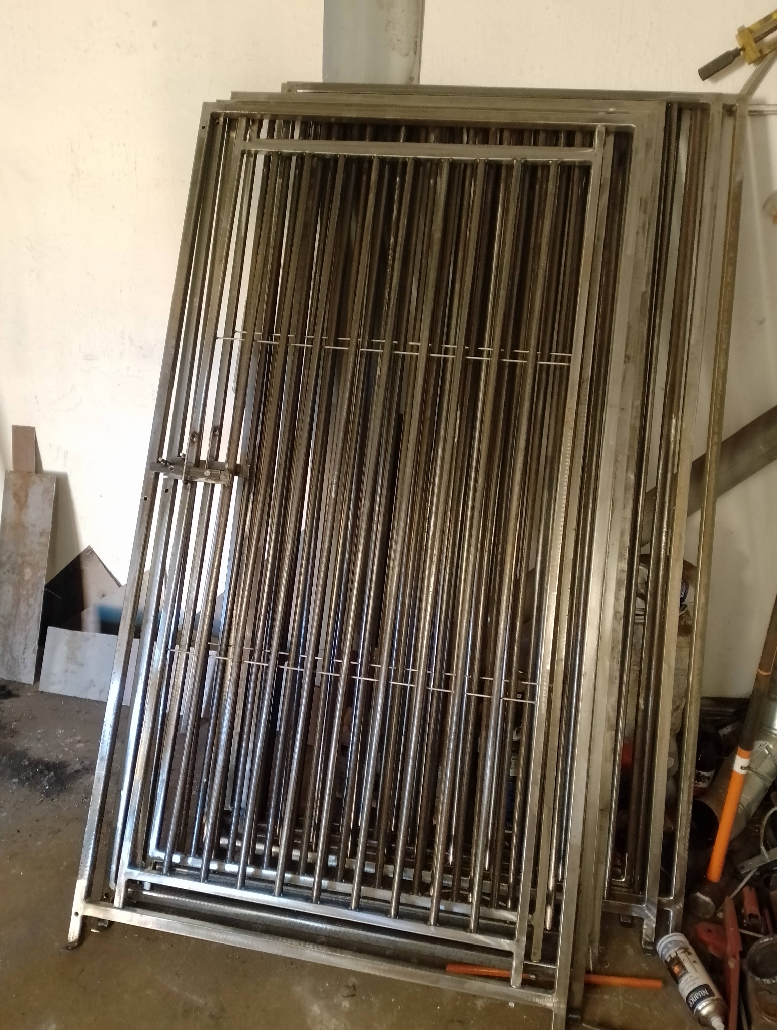 Stos metalowych furtek z zamkiem, wykonanych z rurek i profili, opartych o ścianę w warsztacie. Widoczne ślady obróbki i narzędzia.