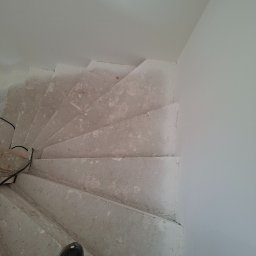 Leszbud - Betonowe schody spiralne w trakcie prac wykończeniowych, widok z góry, fragment stopy w obuwiu roboczym w dolnej części kadru. Surowe ściany w tle.