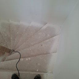 Leszbud - Betonowe schody spiralne w trakcie prac wykończeniowych, widok z góry na stopy w obuwiu roboczym i kabel narzędzia. Surowe wykończenie ścian.