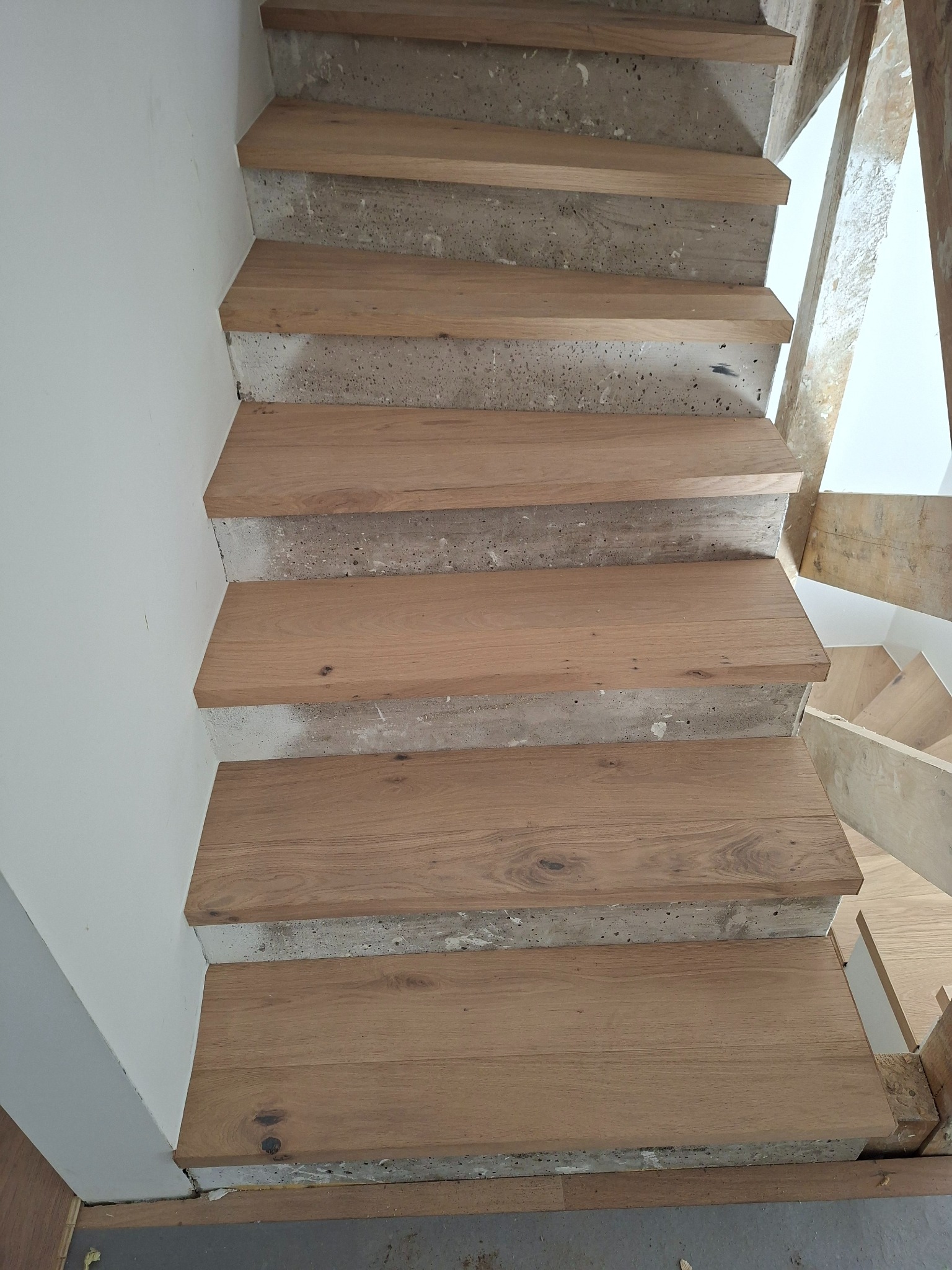 Drewniane stopnie schodów w trakcie montażu, widoczne surowe betonowe podłoże i fragment ściany. Ujęcie z góry, ukazujące detale wykończenia.