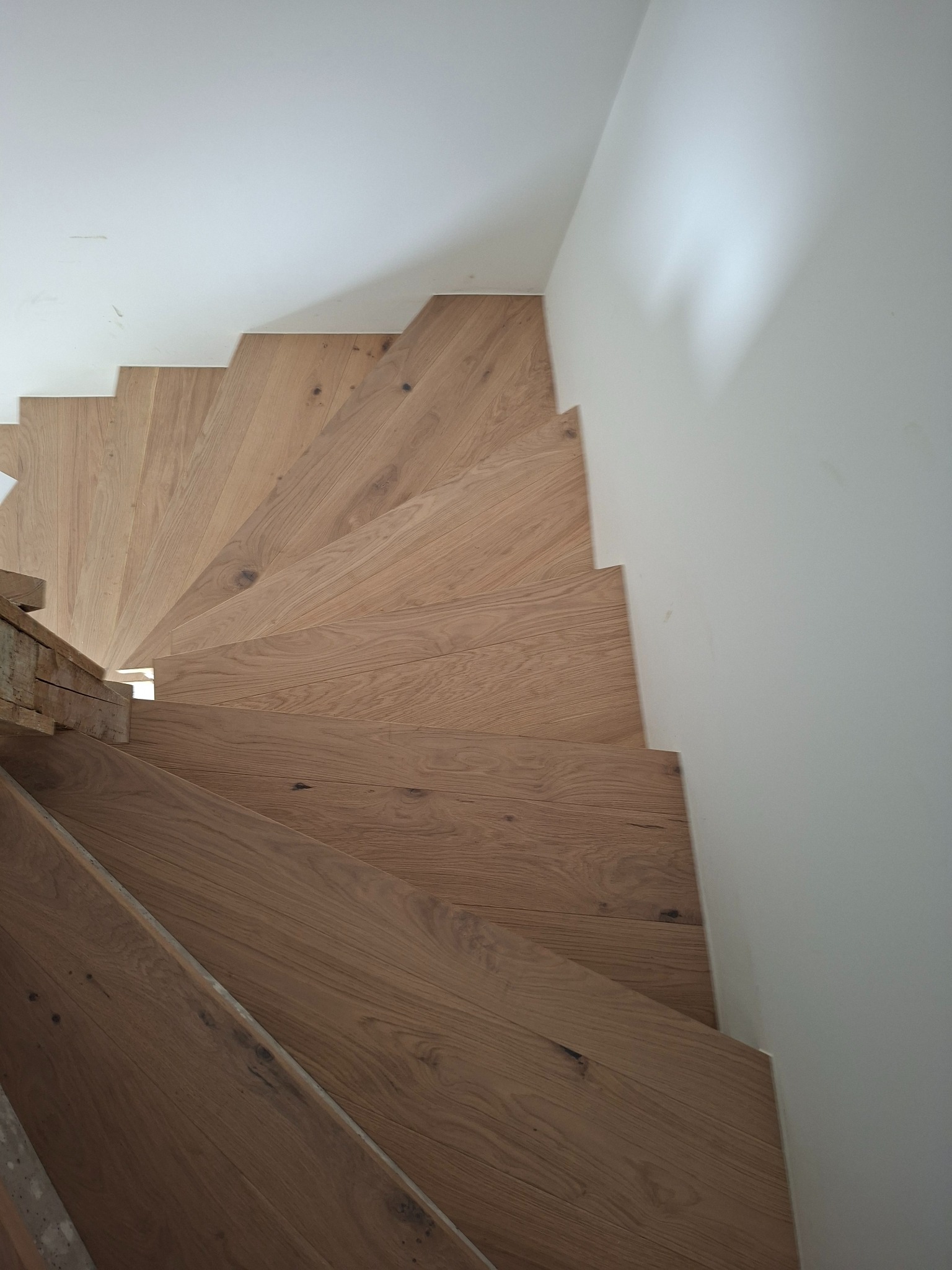 Drewniane schody spiralne z jasnego drewna, widok z góry, z białymi ścianami. Naturalne usłojenie drewna, schody w trakcie montażu, widoczne niedoskonałości wykończenia.