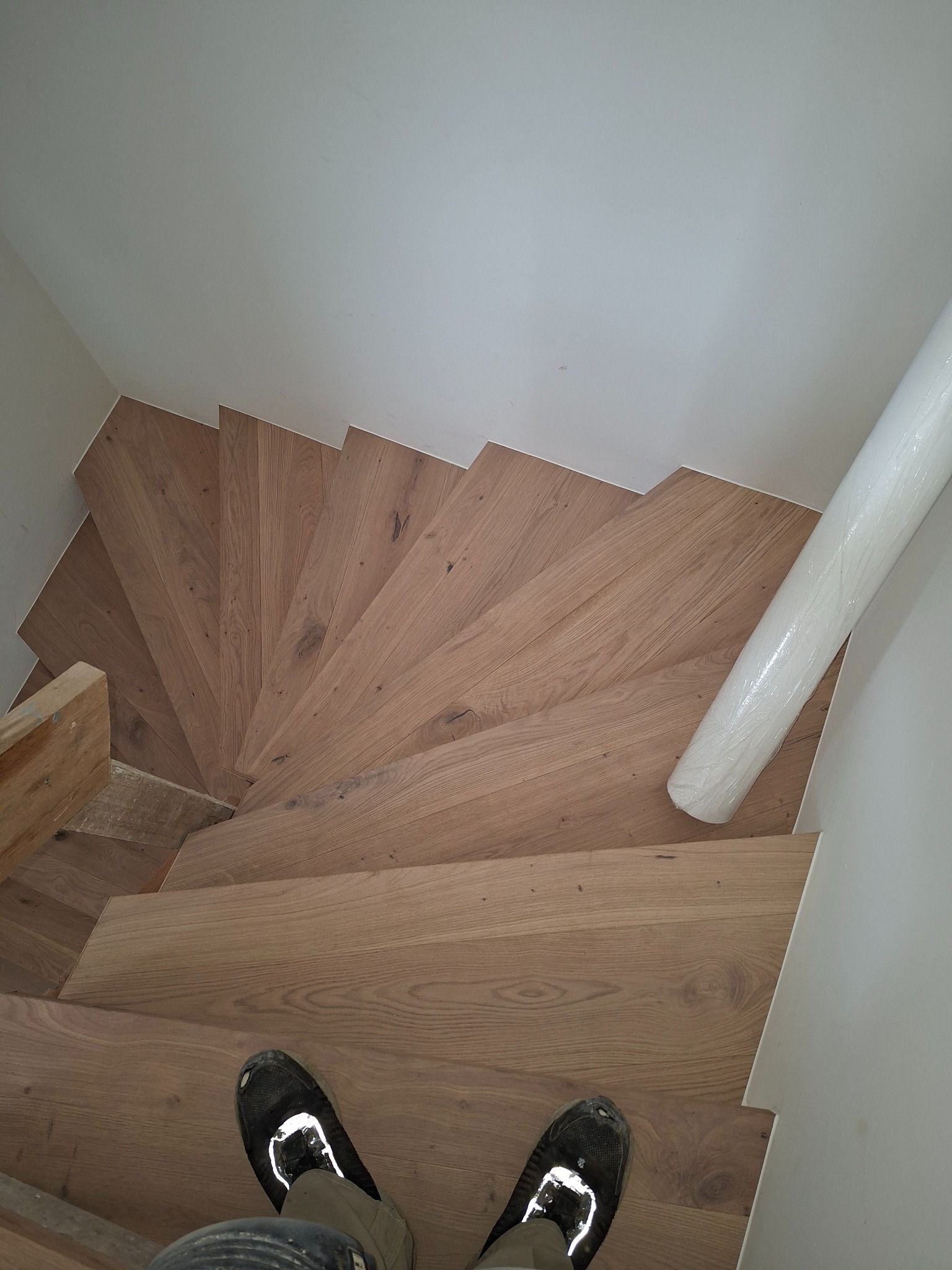 Spiralne schody z jasnego drewna, widok z góry, z butami osoby stojącej na schodach i rolką folii ochronnej przy ścianie. Naturalne oświetlenie.