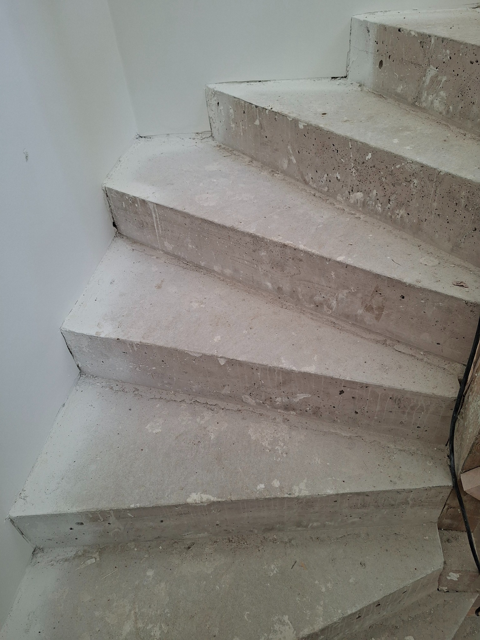 Surowe, betonowe schody spiralne w trakcie budowy, widoczne z góry, z fragmentem białej ściany i kablami w tle. Minimalistyczny, industrialny styl.