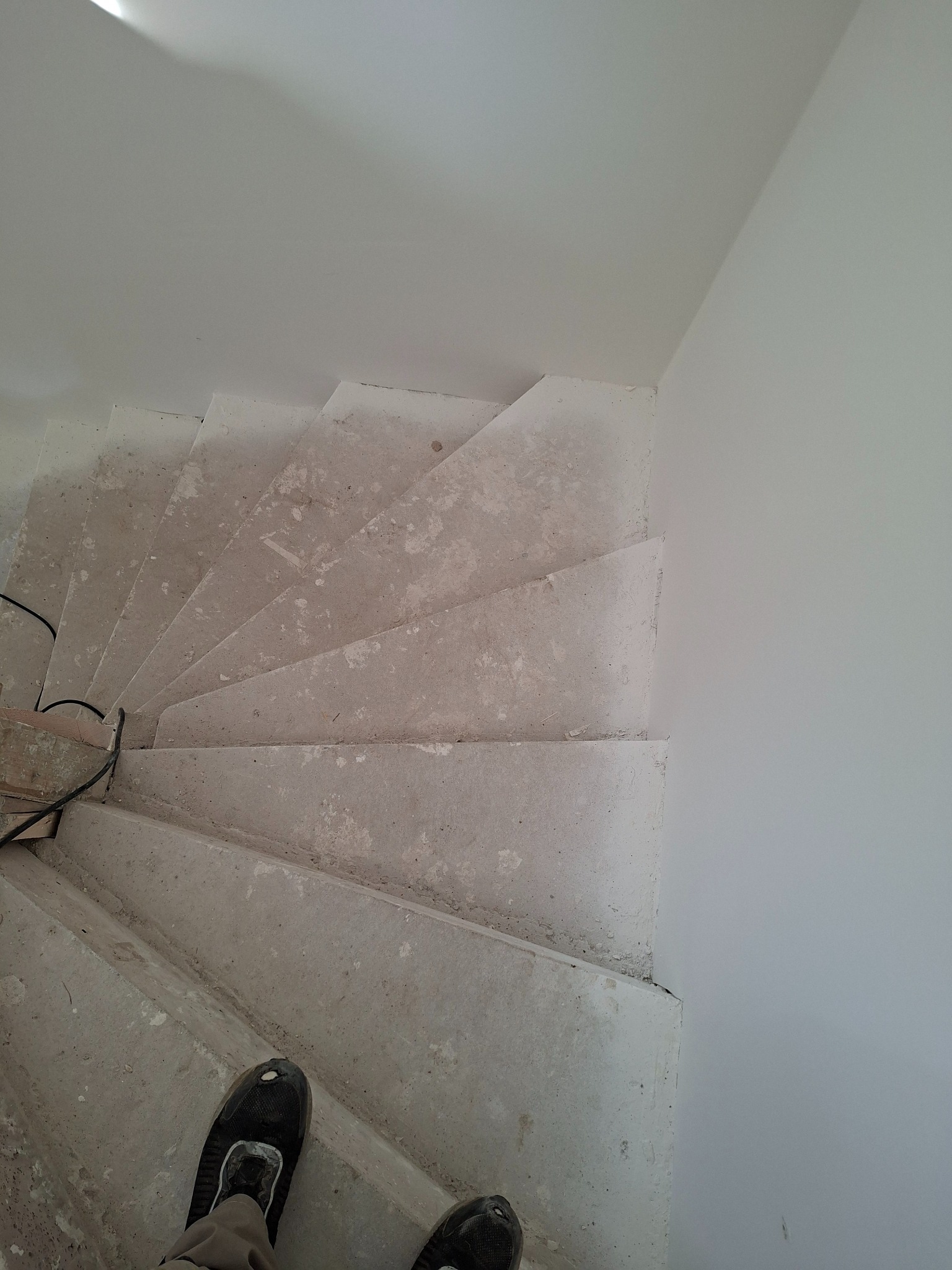 Betonowe schody spiralne w trakcie prac wykończeniowych, widok z góry, fragment stopy w obuwiu roboczym w dolnej części kadru. Surowe ściany w tle.