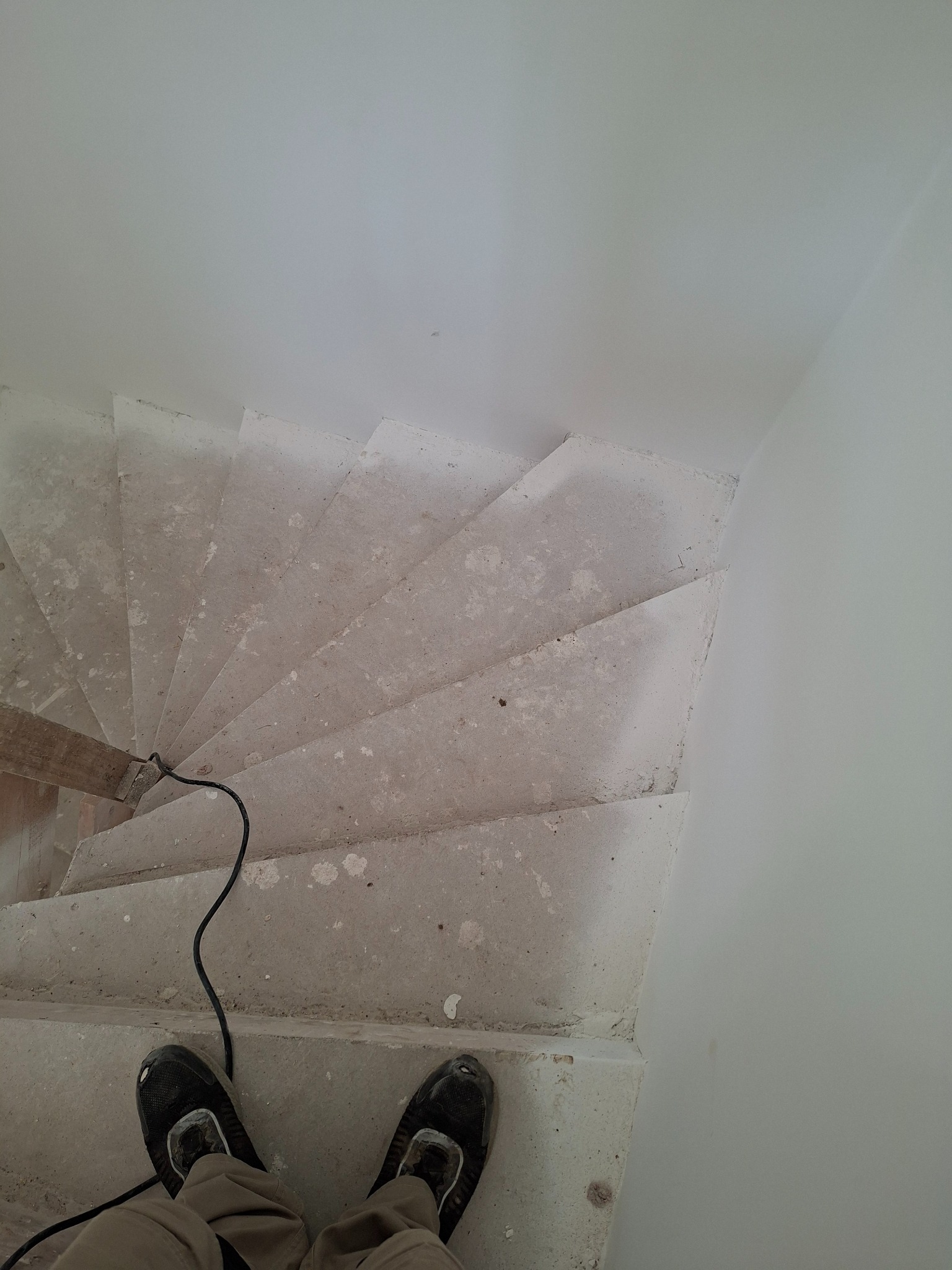Betonowe schody spiralne w trakcie prac wykończeniowych, widok z góry na stopy w obuwiu roboczym i kabel narzędzia. Surowe wykończenie ścian.