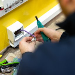 HELPO Network Paweł Surmacz - Instalator łączy przewody w gniazdku sieciowym na żółtej ścianie, używając narzędzia do zaciskania. Widoczne kable, gniazdko i fragment narzędzia.