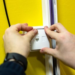 HELPO Network Paweł Surmacz - Montaż gniazda sieciowego RJ45 na żółtej ścianie, widoczne ręce instalatora, listwa przypodłogowa z kablem. Instalacja sieci LAN w biurze lub domu.