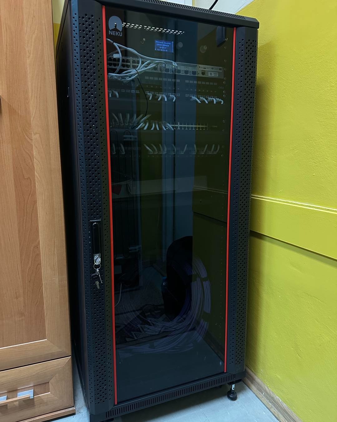 Czarna szafa rack z oszklonymi drzwiami, widoczne switche i okablowanie, umieszczona w pomieszczeniu z żółtą ścianą. Klucze w zamku szafy.