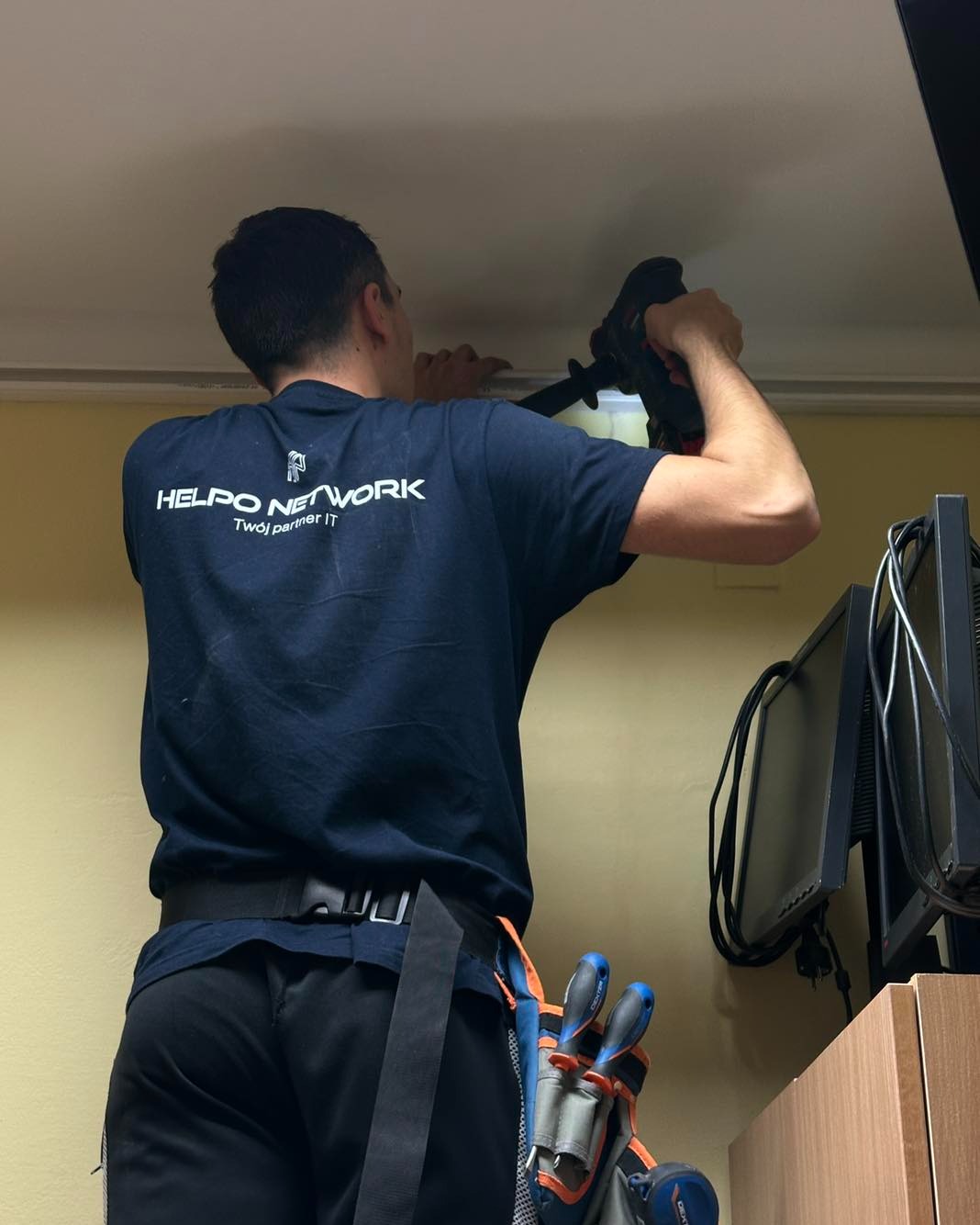Pracownik firmy IT montuje element pod sufitem biura, ubrany w firmową koszulkę z logo 'Helpo Network'. Widoczne narzędzia i monitory komputerowe w tle.