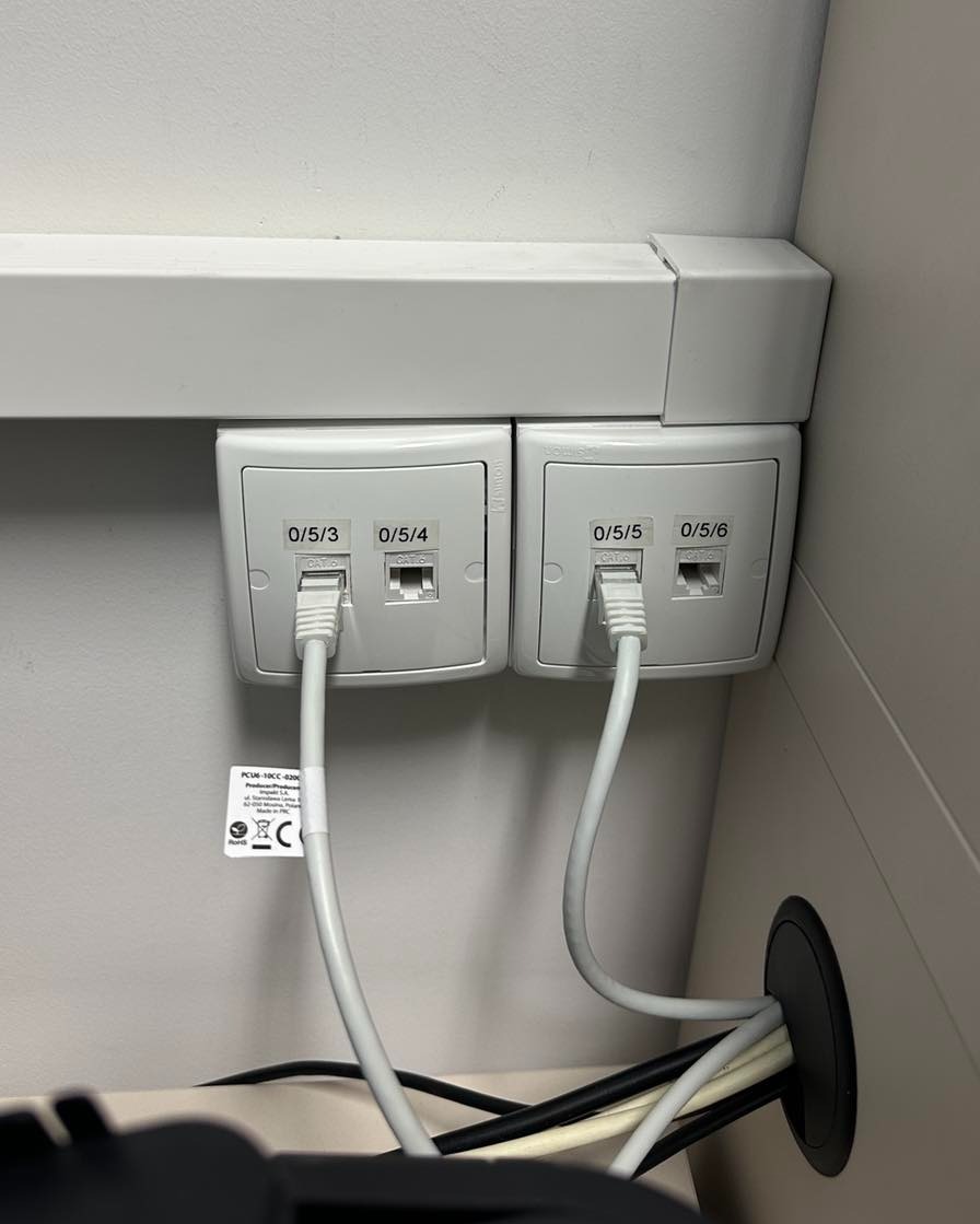 Dwa podwójne gniazda sieciowe RJ45 z podłączonymi kablami, oznaczone numerami 0/5/3, 0/5/4, 0/5/5 i 0/5/6, zamontowane na ścianie w biurze. Widoczne kable w przepuście.