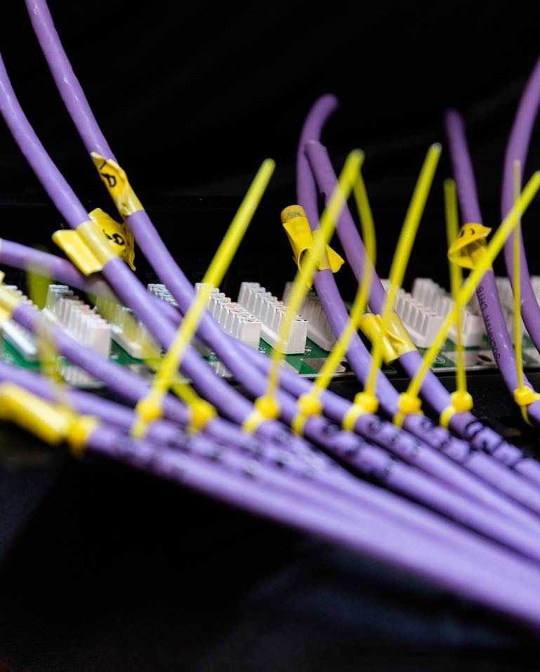 Fioletowe kable sieciowe z żółtymi opaskami zaciskowymi, podłączone do białych portów na zielonej płycie. Widoczne oznaczenia na kablach.