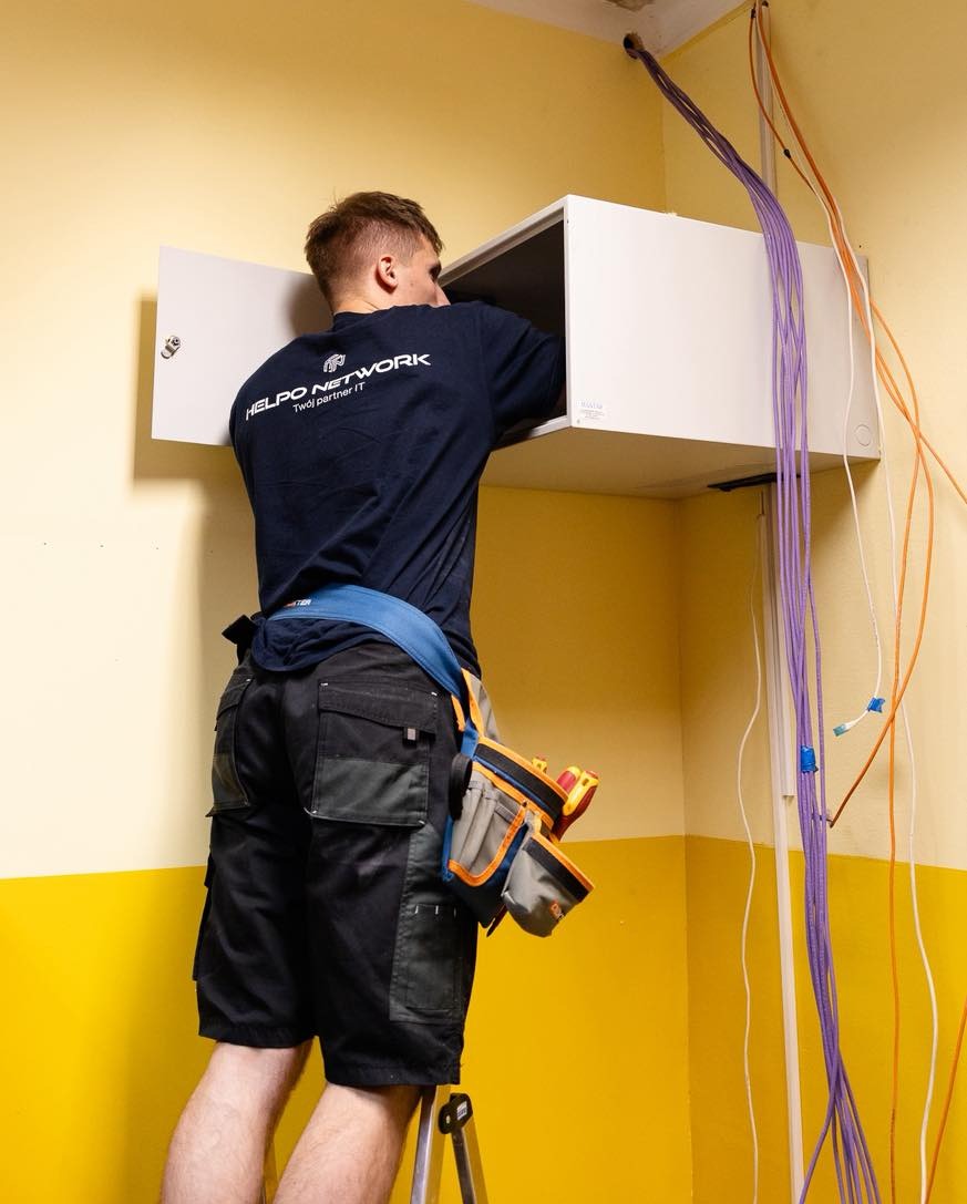 Instalator w firmowej koszulce montuje szafkę z kablami sieciowymi w rogu pomieszczenia o żółtych ścianach. Widoczne narzędzia i kable w kolorze pomarańczowym i fioletowym.