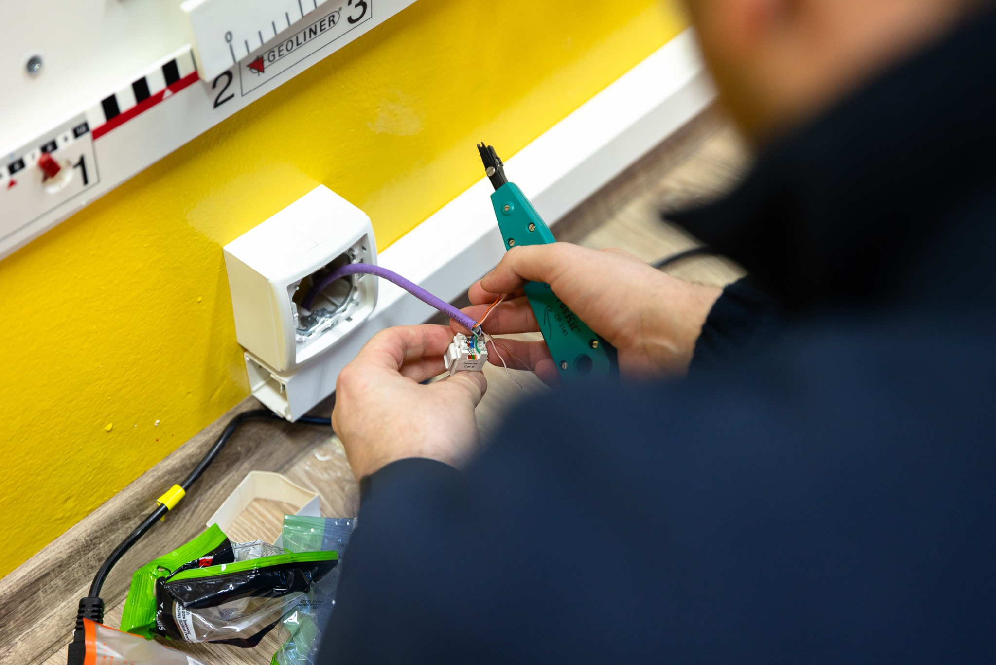 Instalator łączy przewody w gniazdku sieciowym na żółtej ścianie, używając narzędzia do zaciskania. Widoczne kable, gniazdko i fragment narzędzia.